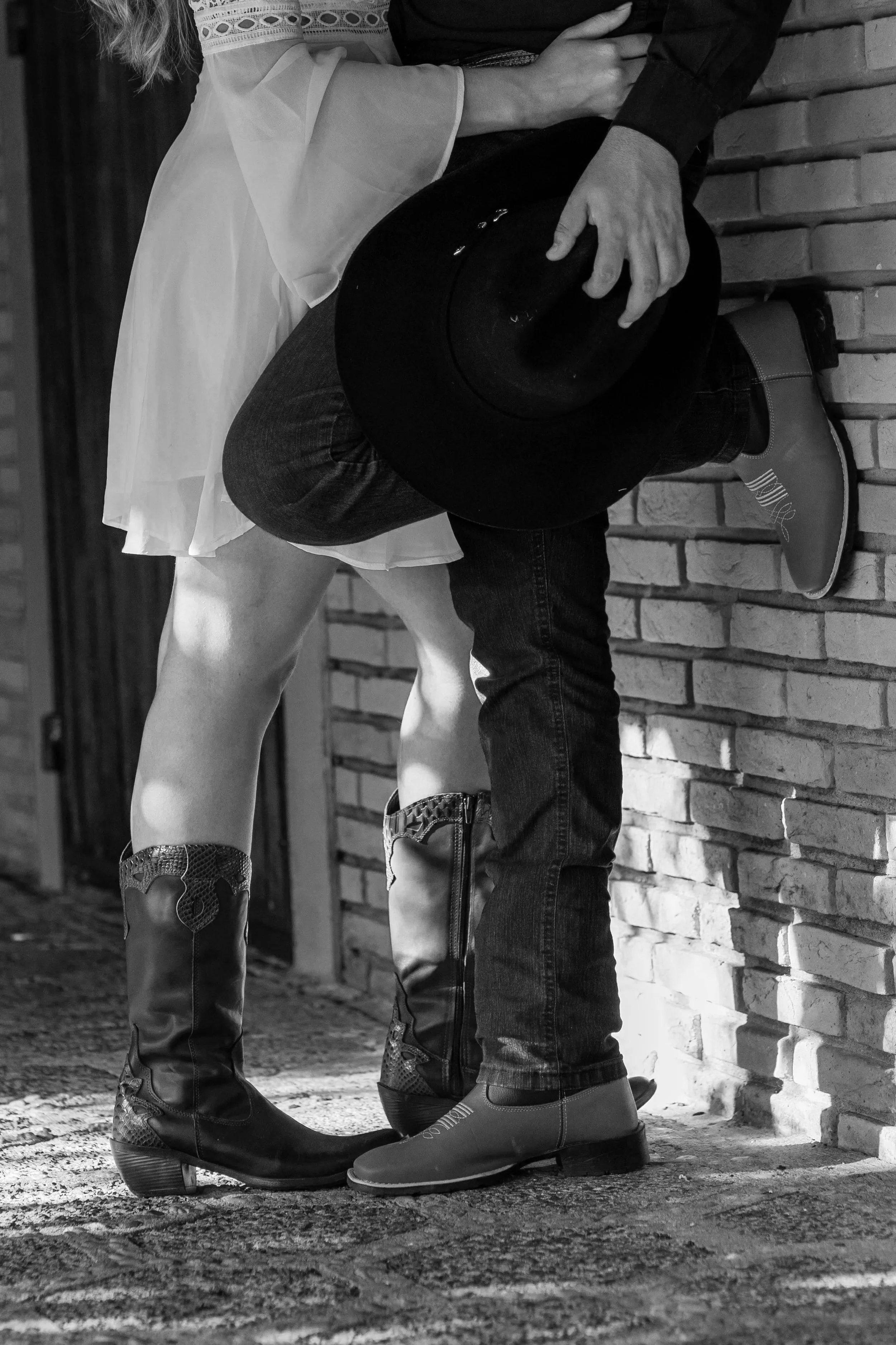 A person wearing cowboy boots, jeans, and a white skirt standing close to a person in dark jeans, a dark shirt, and cowboy boots, holding a hat, against a brick wall.