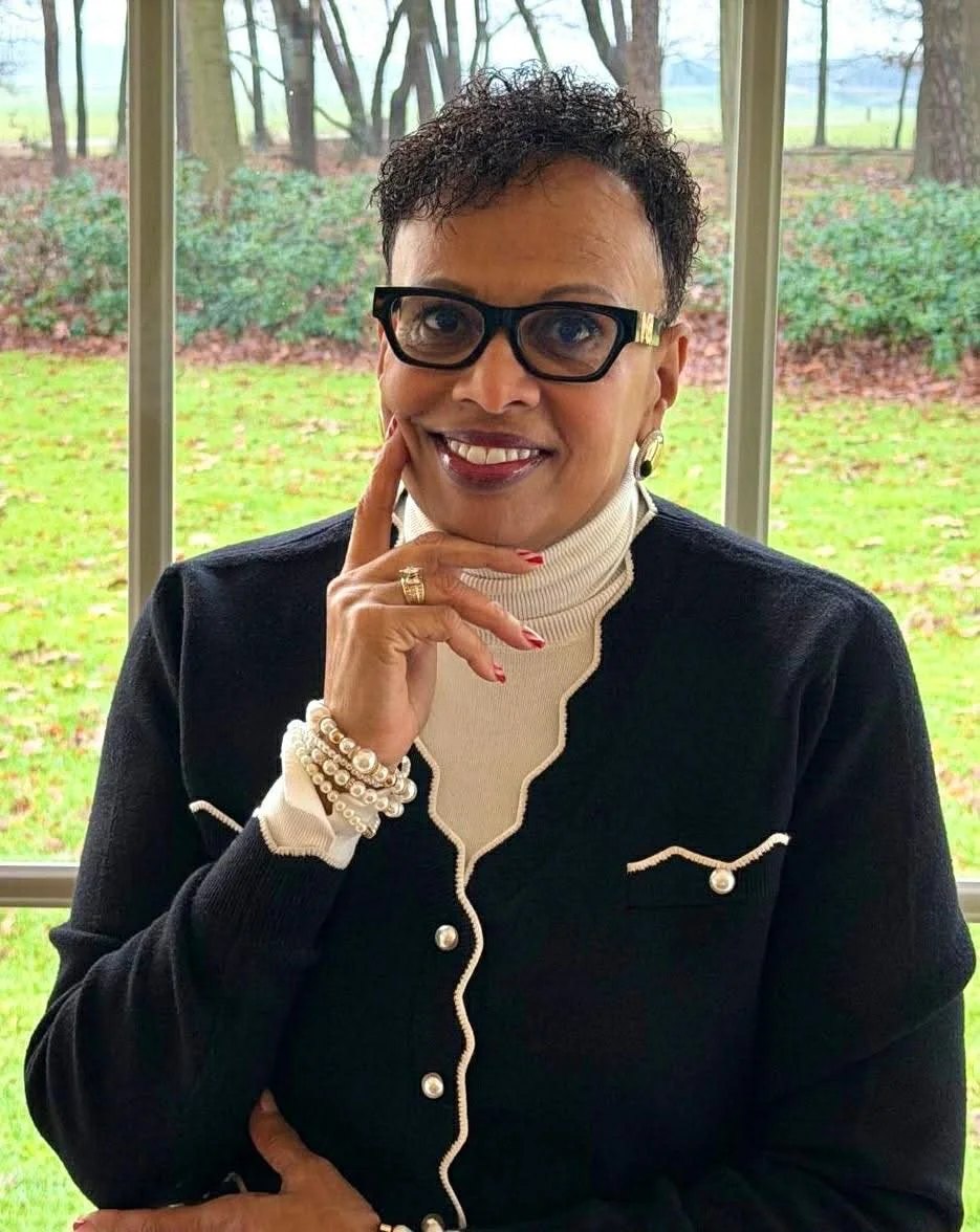A woman with short curly hair, glasses, and earrings smiling, wearing a red quilted jacket and black turtleneck in front of a bookshelf filled with colorful books.
