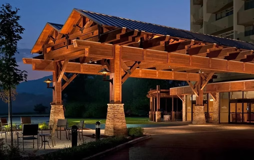 Exterior view of a hotel entrance with a large wooden canopy supported by stone pillars, outdoor seating, and ambient lighting during dusk.