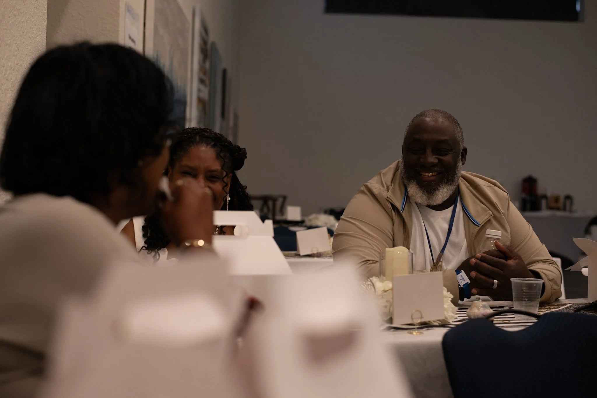 Three people sitting at a table, engaging in conversation and smiling, in a dimly lit indoor setting.