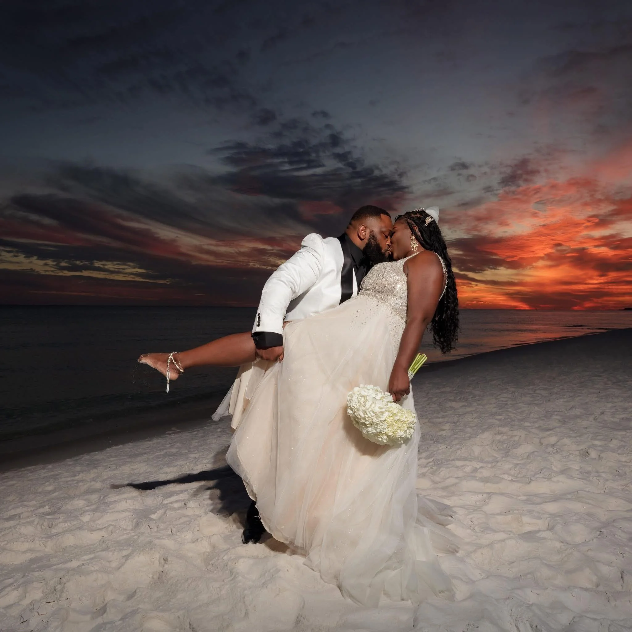 A couple dressed in wedding attire on a beach at sunset, with the man in a white tuxedo lifting the woman, who is holding a bouquet of white flowers, as they kiss.