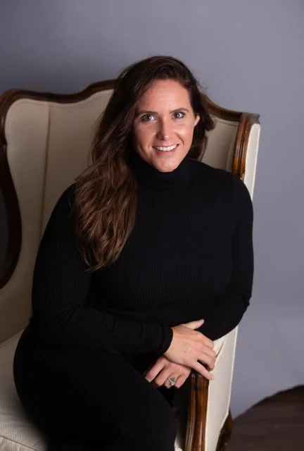 A woman with long brown hair, wearing a black turtleneck sweater, sitting on a vintage-style cream-colored armchair with wooden accents, smiling at the camera.