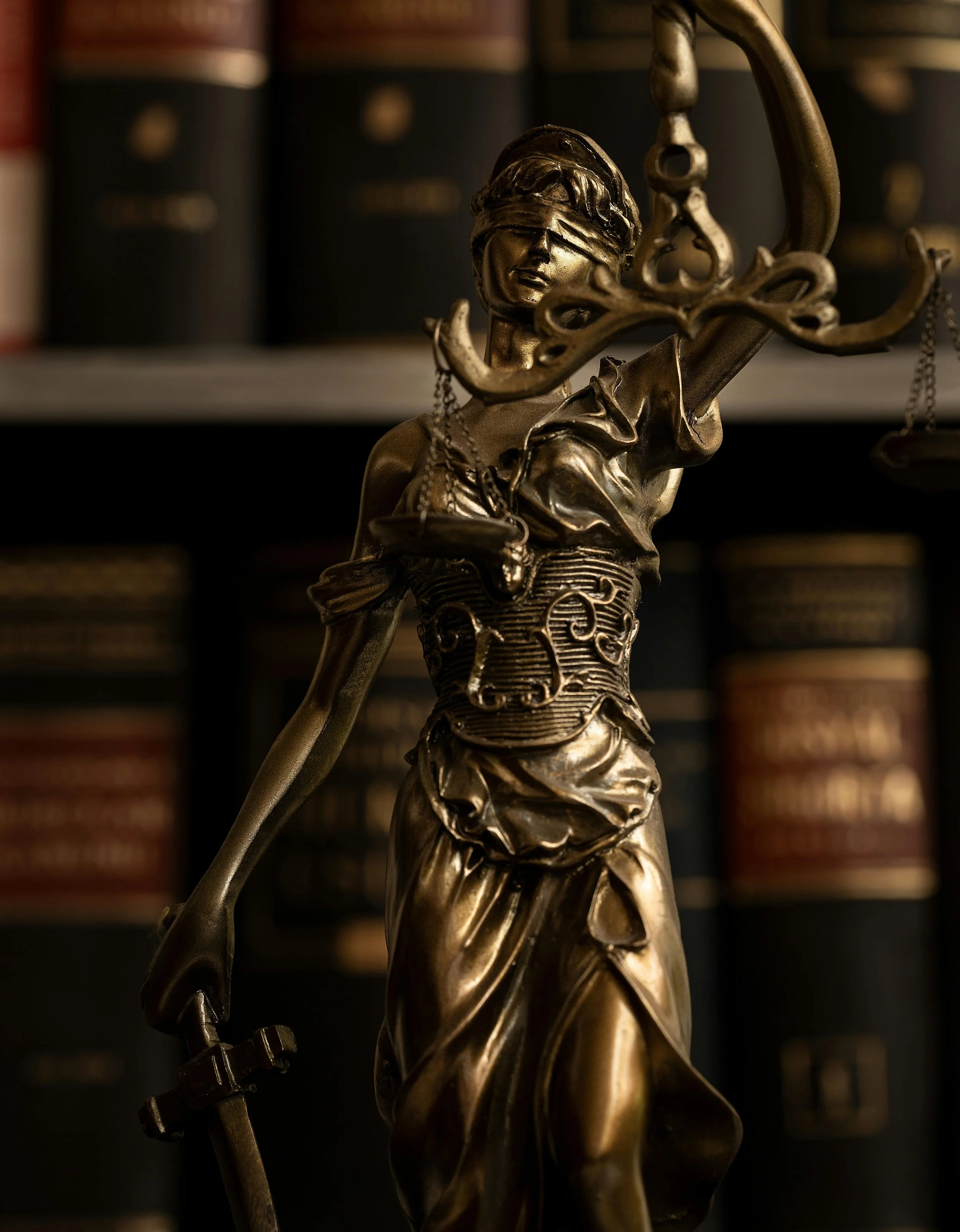 A bronze statue of Lady Justice with a blindfold, holding scales in her left hand and a sword in her right hand, standing in front of a bookshelf filled with legal books.