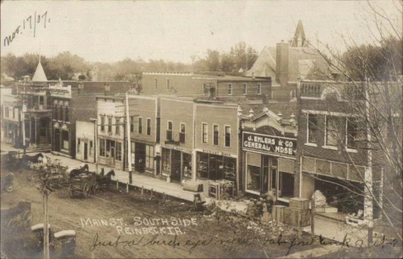 Historical black and white photo of a Main Street South Side in Reinbeck, showing brick buildings with storefronts, horse-drawn carriages parked along the street, and handwritten notes indicating the date March 17, 1907, and location details.