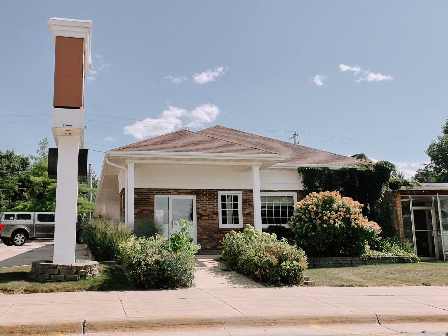 A small commercial building with a brick facade and white trim, surrounded by garden bushes, with a parking lot and a silver truck on the left, and a signpost with a blank sign on the sidewalk in front, under a partly cloudy sky.