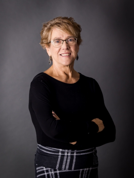 A woman with short blonde hair, glasses, and earrings, wearing a black long-sleeve shirt and a plaid skirt, standing against a gray background with arms crossed and smiling at the camera.