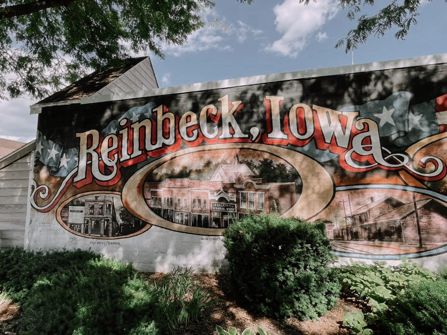 Large vintage-style sign that reads 'Reinbeck, Iowa' with illustrations of old buildings and streets of Reinbeck, Iowa, surrounded by green bushes and trees under a partly cloudy sky.
