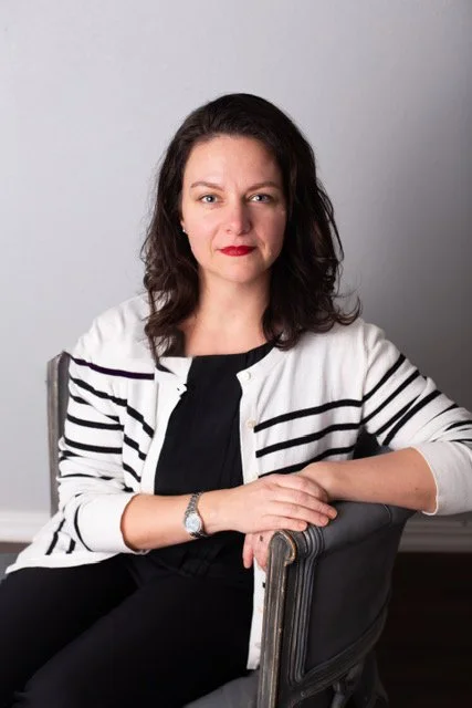 A woman with shoulder-length dark hair, red lipstick, and fair skin, sitting on a chair against a plain gray wall, wearing a black top and a white cardigan with black stripes.