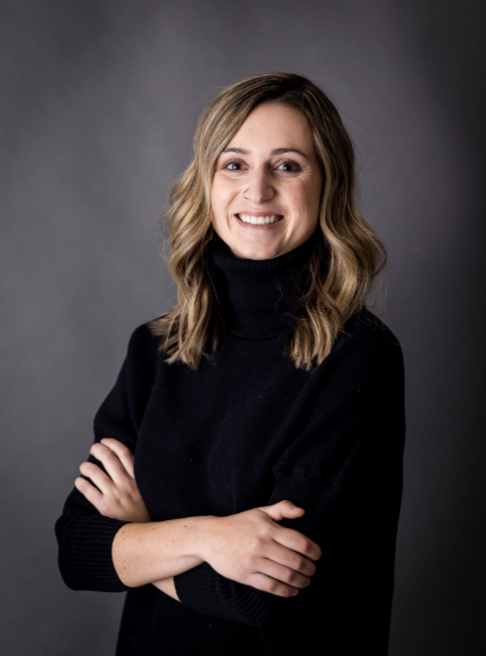 A woman with shoulder-length wavy blonde hair, smiling, wearing a black turtleneck sweater, standing with arms crossed against a gray background.