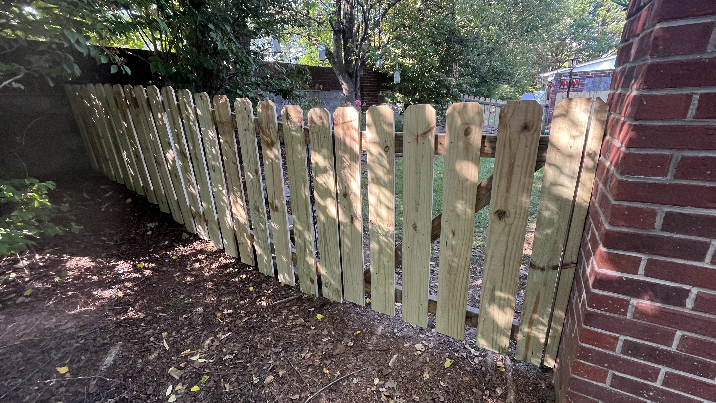 New wooden picket fence installed next to brick wall, with trees and garden in background.