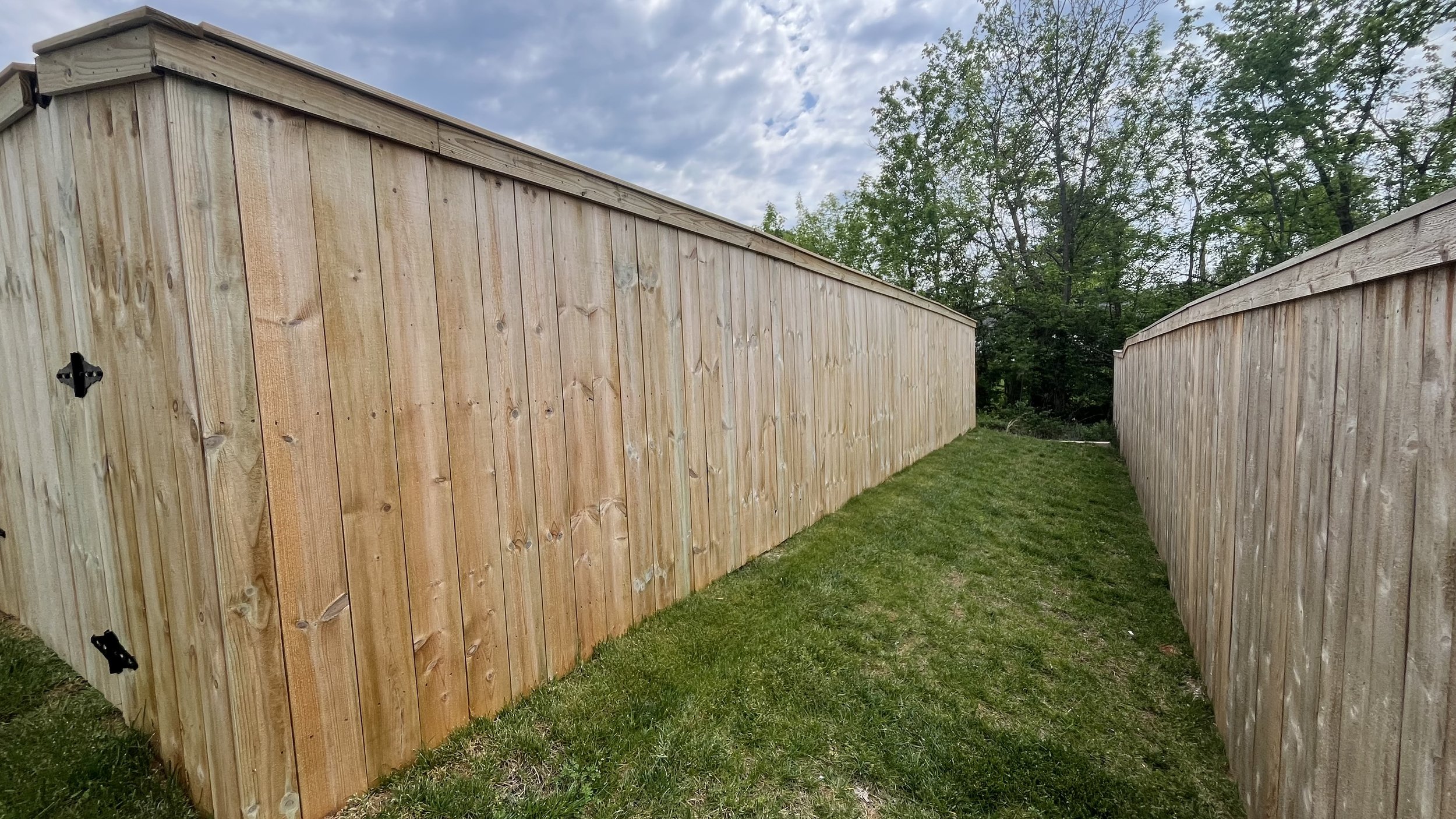 View of two wooden fences with a grassy pathway between them and trees in the background under a partly cloudy sky.