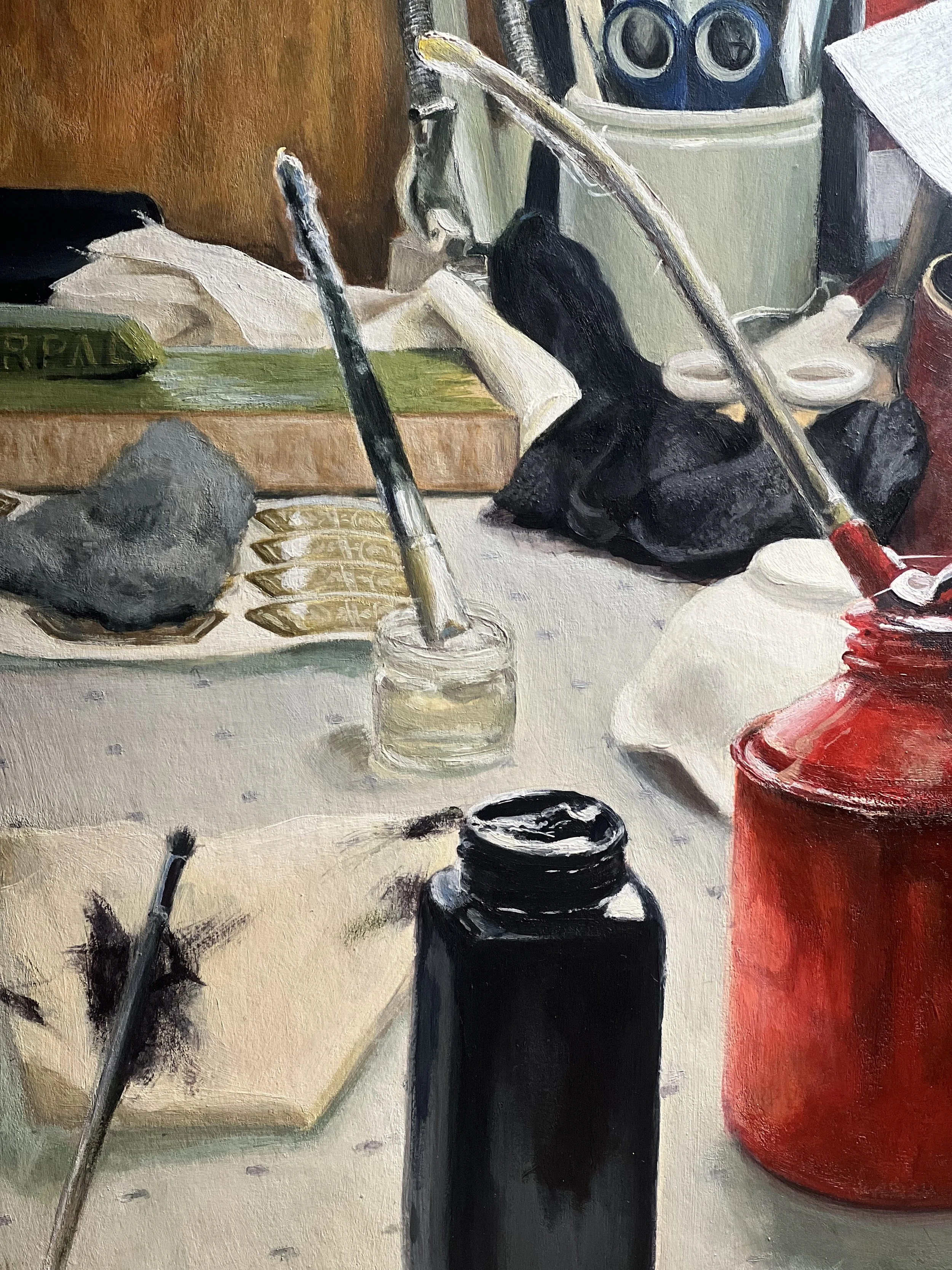 Close-up of an artist's workspace with paint brushes, a paper with sketch marks, ink bottle, and various art supplies on a cluttered table.