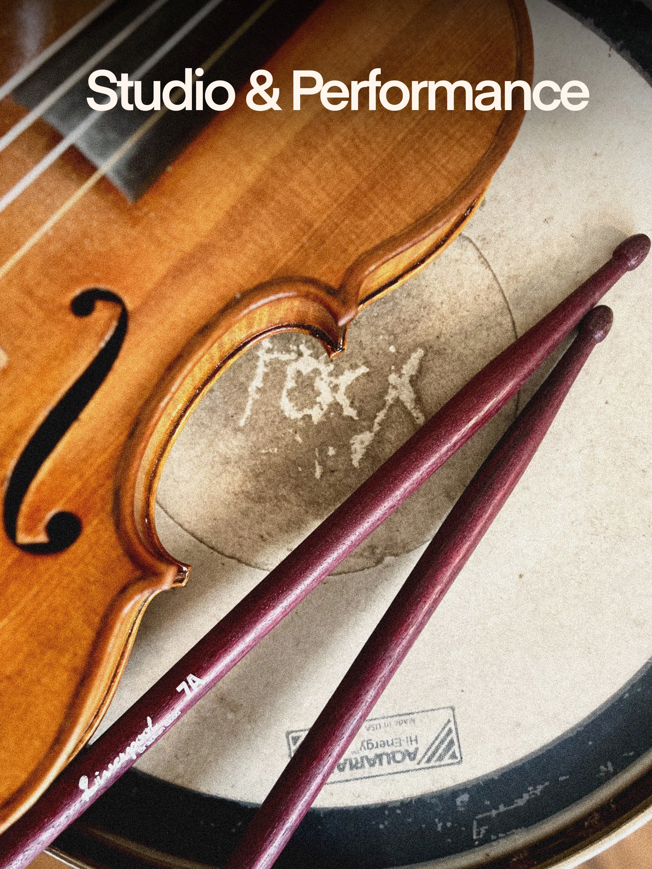 A violin and two bows resting on a large drum with a drumhead marked 'AOLIAN'. The violin's body is visible along with two reddish-purple bows.