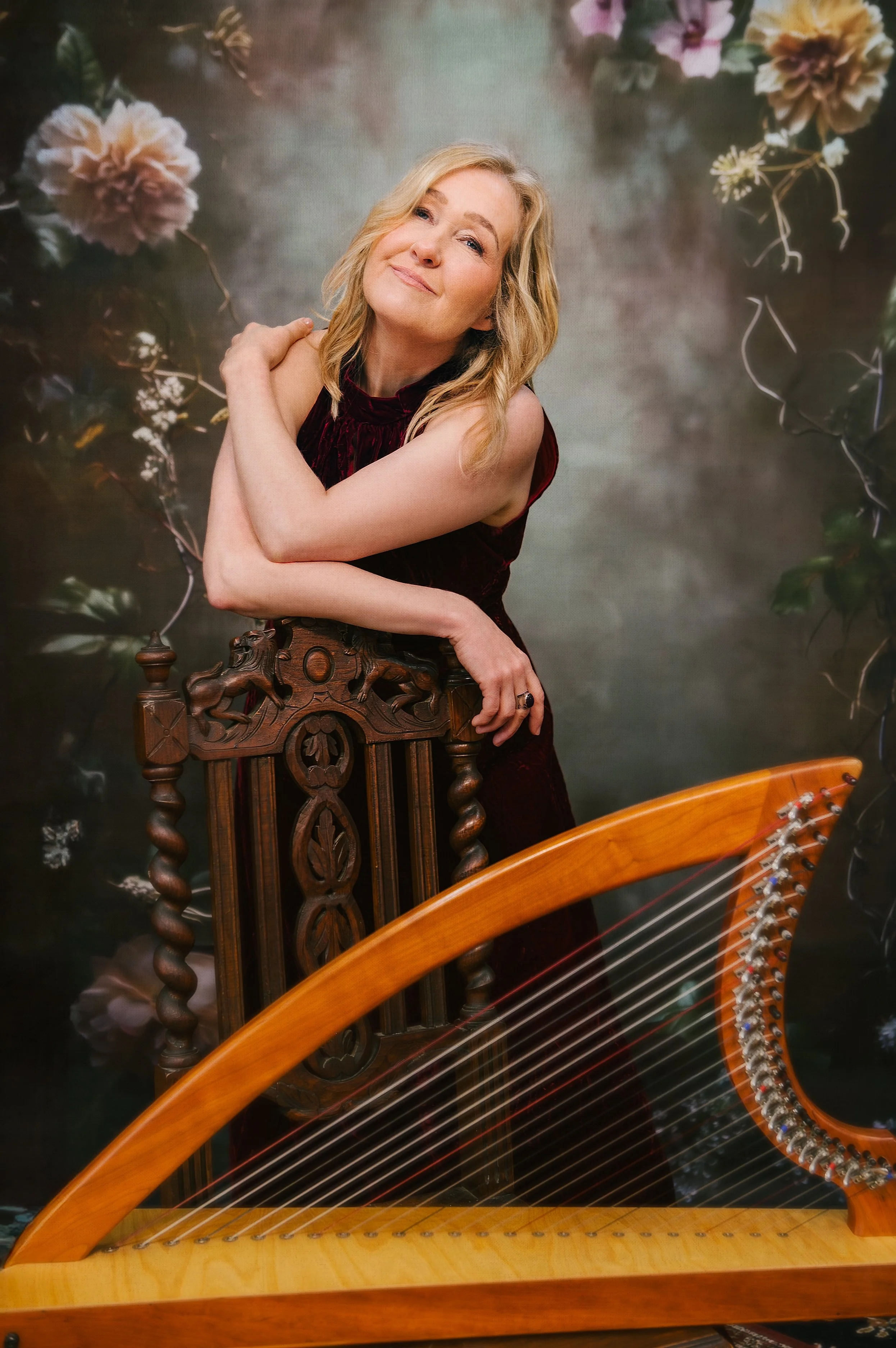 A woman with blonde hair in a burgundy velvet dress, smiling and hugging a wooden carved chair, standing behind a harp with a floral backdrop.