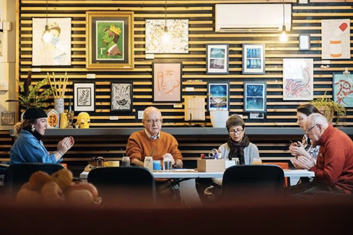 Five people sitting at a table in an art gallery or cafe, praying or meditating with their hands together, with colorful artwork and plants on the wall behind them.