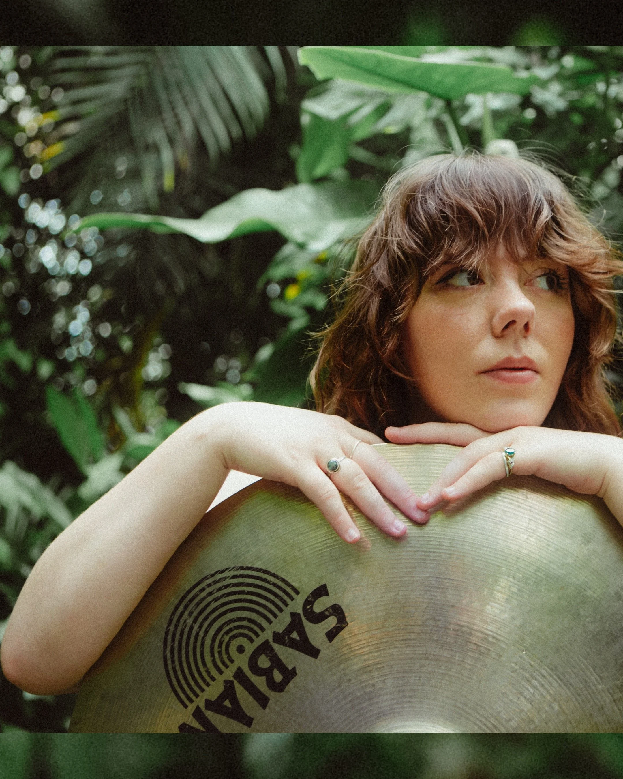 A photo of Meg with wavy brown hair, resting her chin and arms on a Sabian cymbal, with a lush green botanical background.