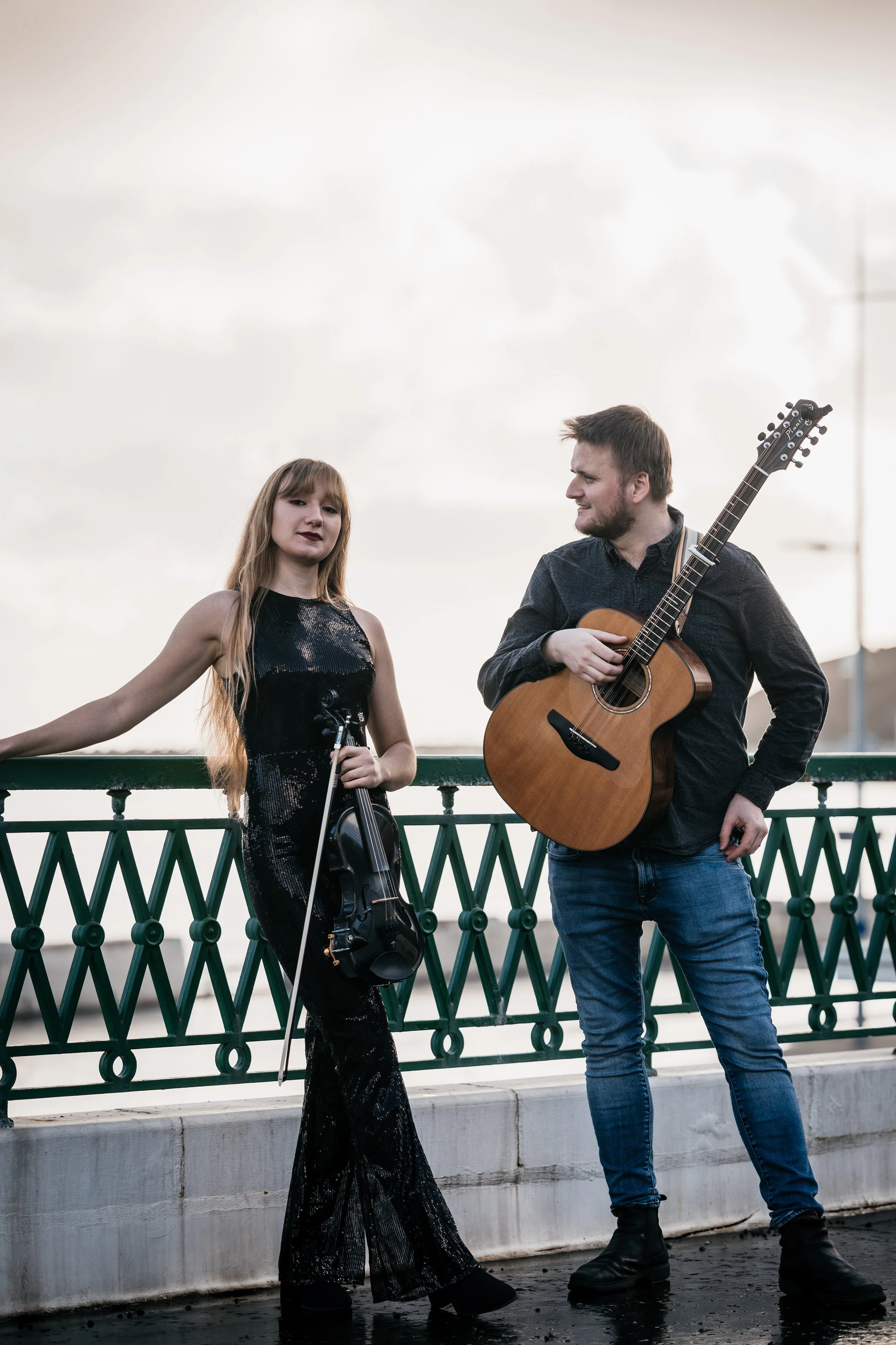 A young woman with a violin and a man with an acoustic guitar standing on a balcony railing, engaging in a musical performance outdoors.