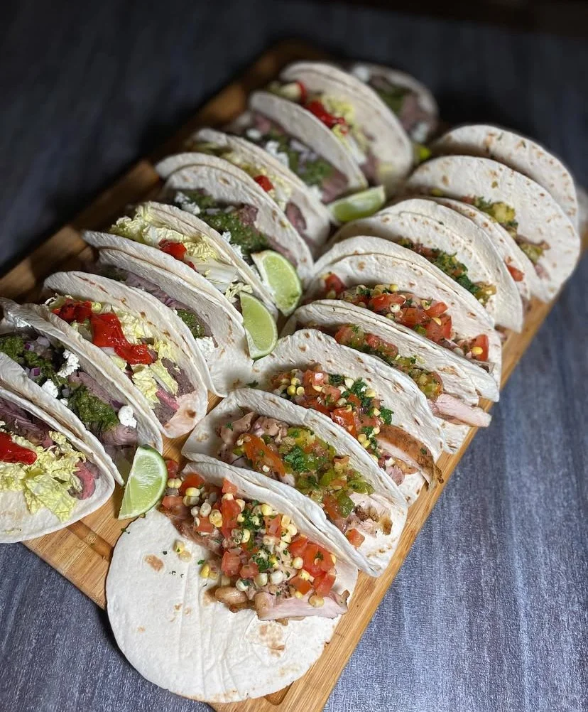Assorted tacos with vegetables, cilantro, diced tomatoes, and lime wedges on a wooden platter.