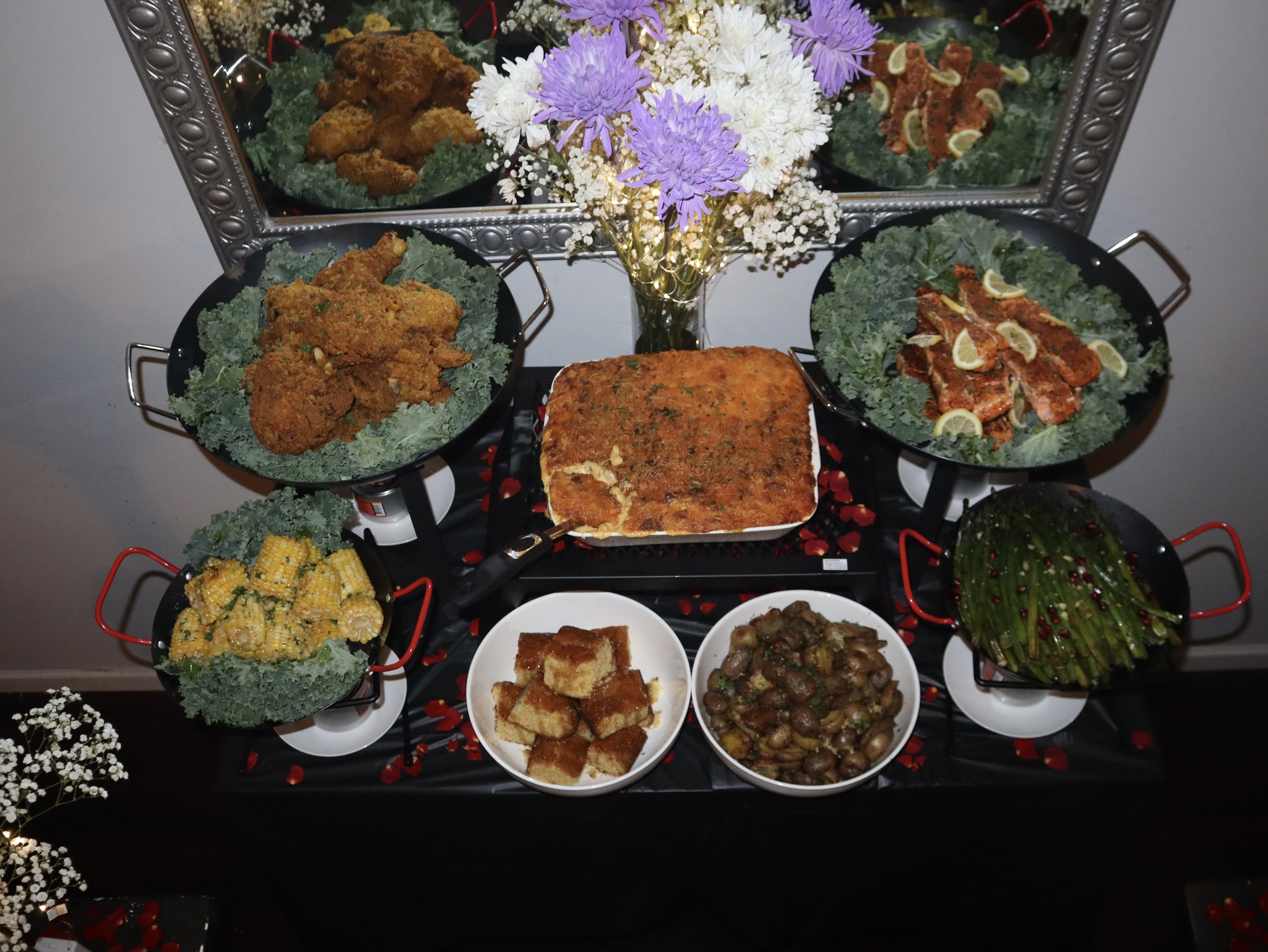 A festive dinner table with fried chicken, lasagna, grilled salmon with lemon slices, corn on the cob, sautéed mushrooms, green beans, and a vegetable side dish, decorated with flowers and a mirror reflection.