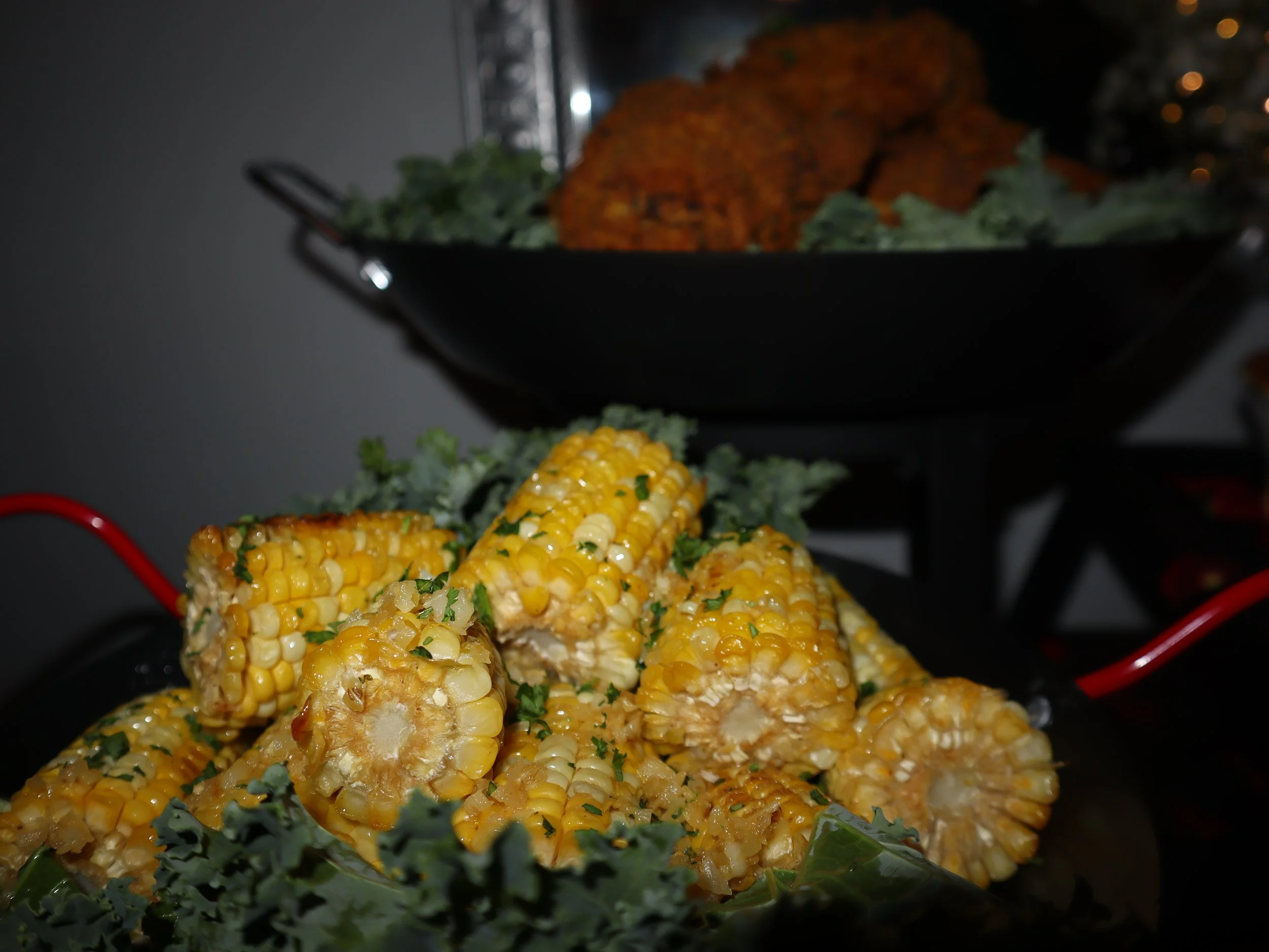 Grilled corn on the cob garnished with herbs, served on a platter with kale, with cooked food in the background.