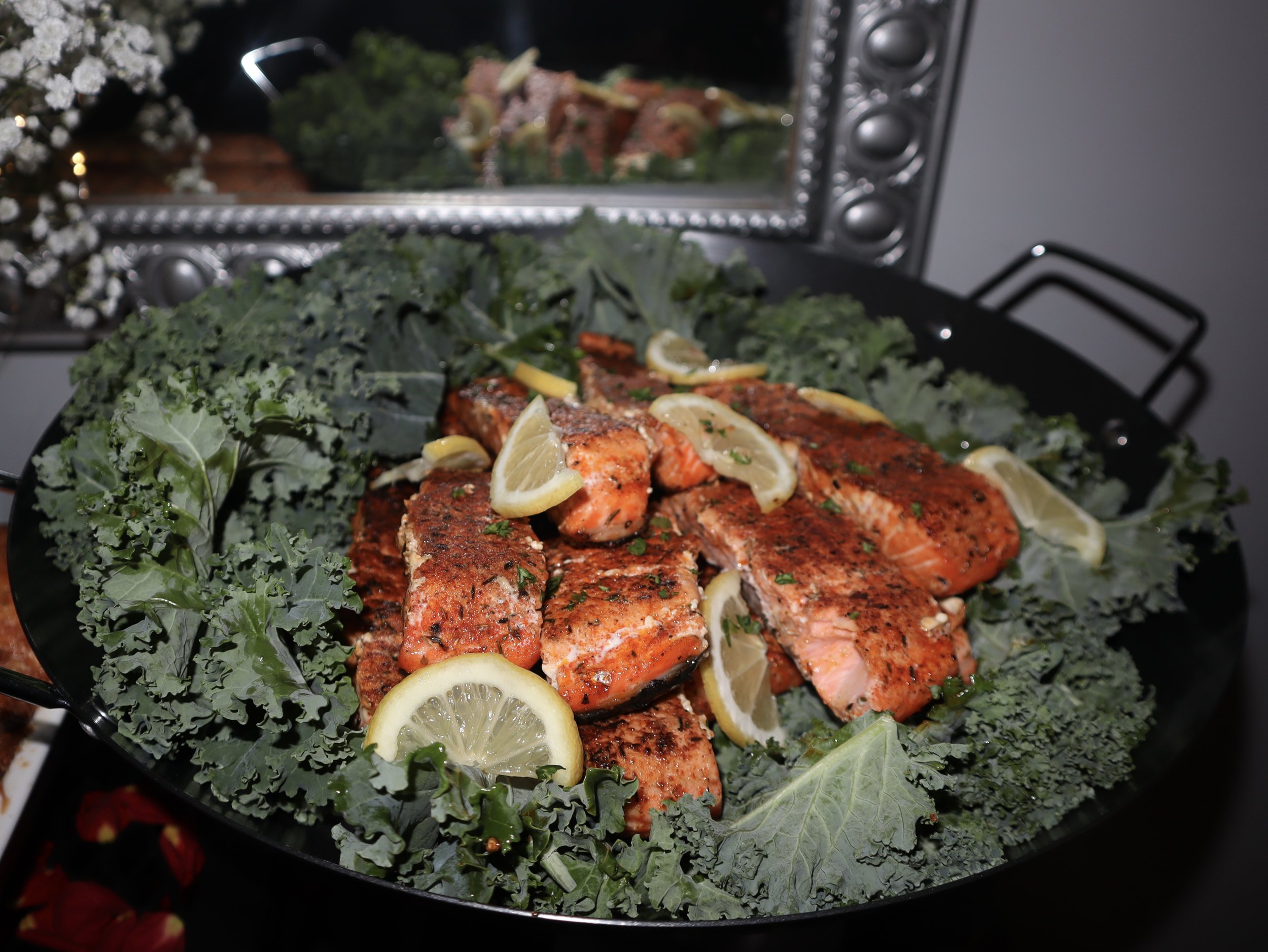 Blackened salmon fillets garnished with lemon slices, placed on a bed of kale greens, in a black serving dish.