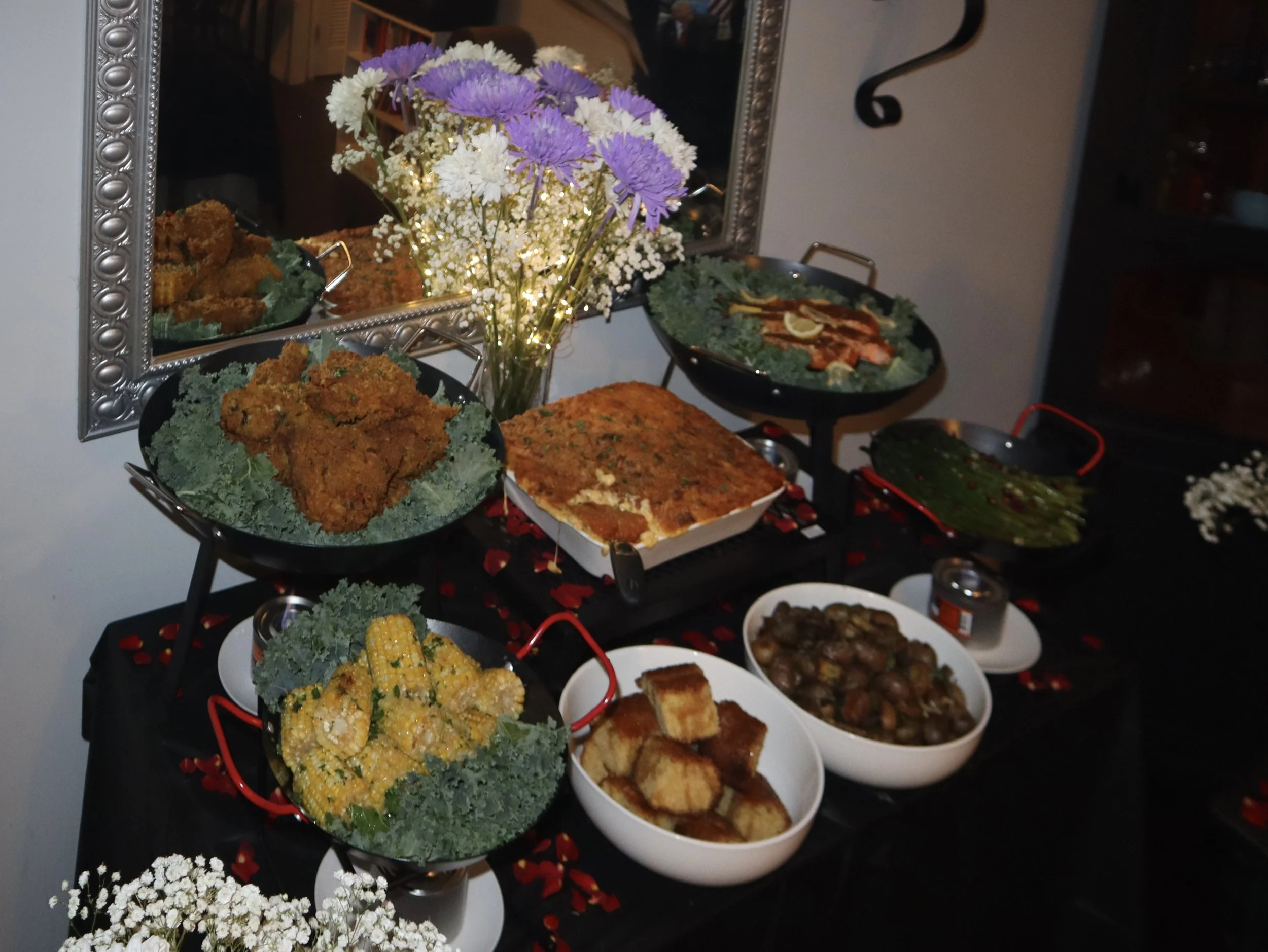 A table with various dishes of fried chicken, baked casserole, green beans, corn, mushrooms, and shrimp, decorated with flowers and rose petals.