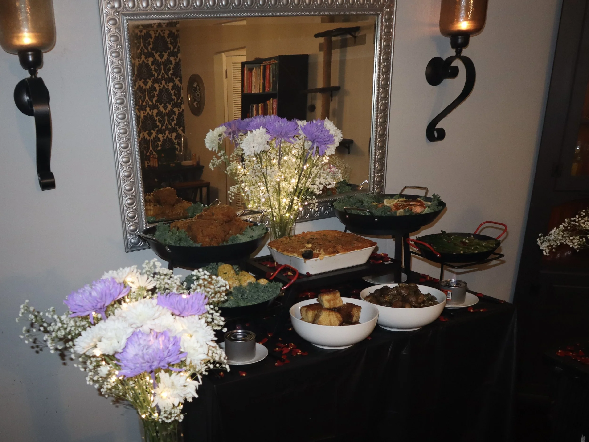 A decorated table with various dishes, bowls of food, and floral arrangements, reflected in a large ornate mirror.