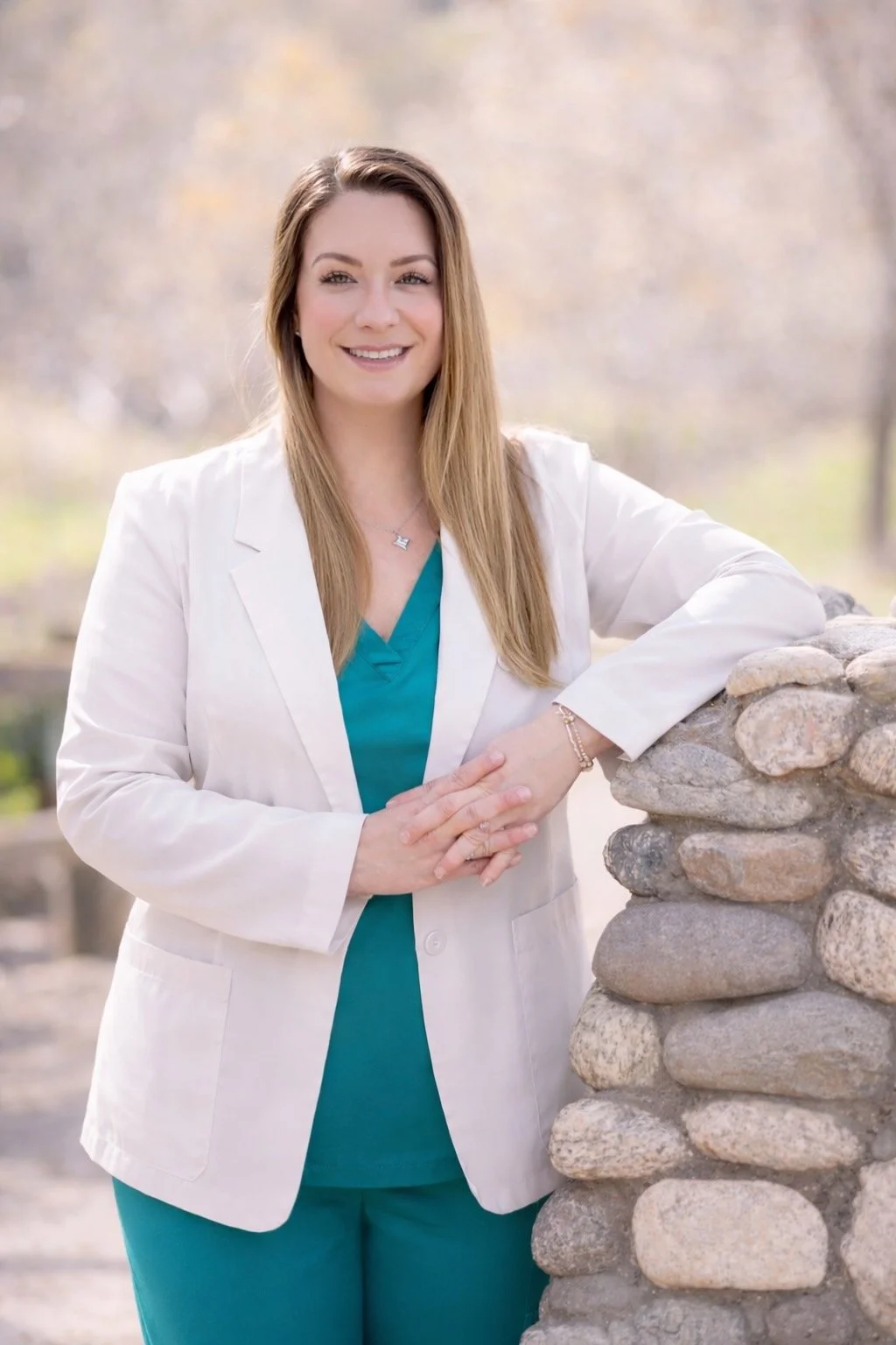 Amy Du, PMHNP-BC in teal scrubs and a white blazer, smiling, leaning on a stone pillar outdoors with blurred trees in the background.