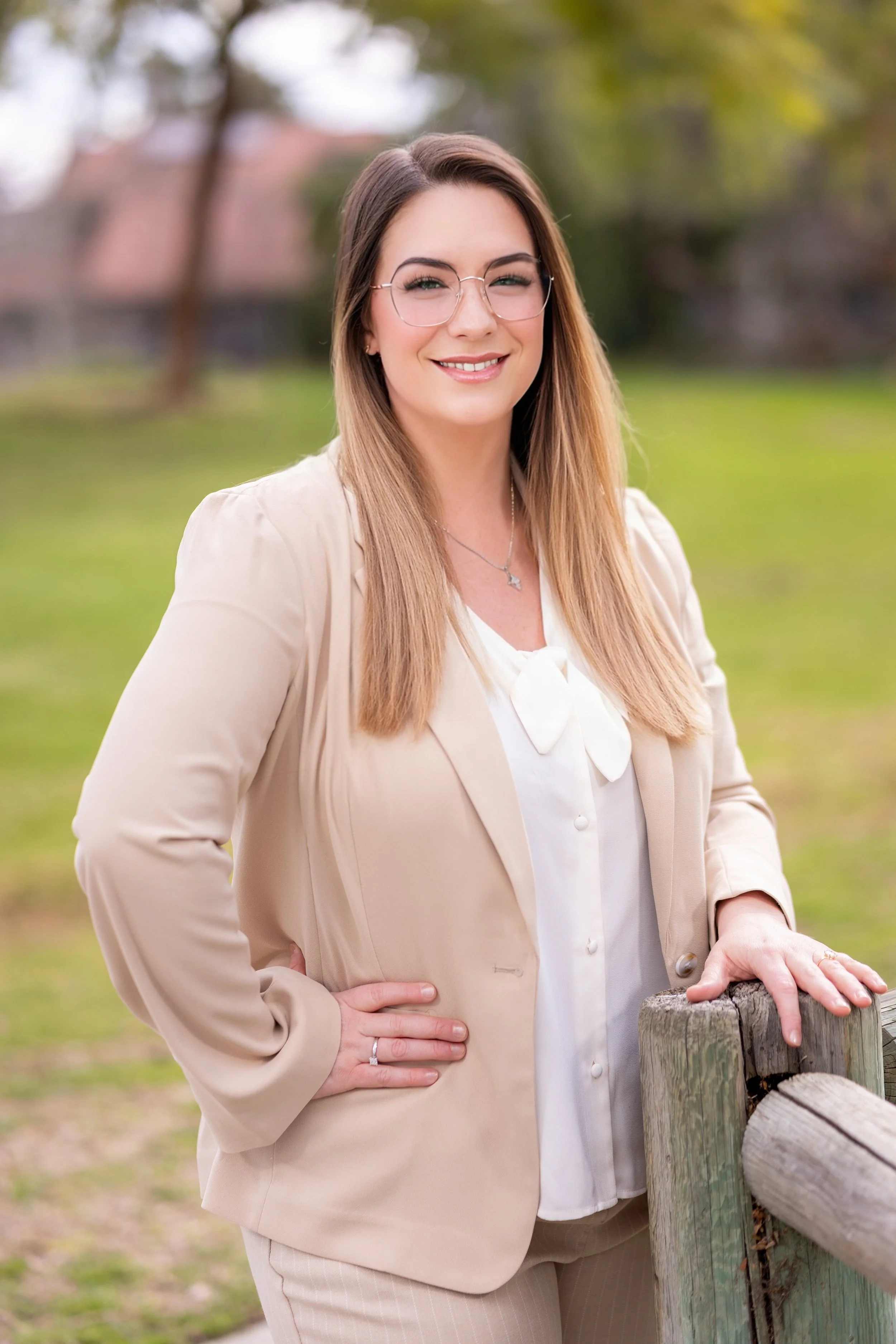 Amy Du, PMHNP, wearing glasses, a beige blazer, and white blouse, smiling outdoors in a park with greenery and trees in the background.
