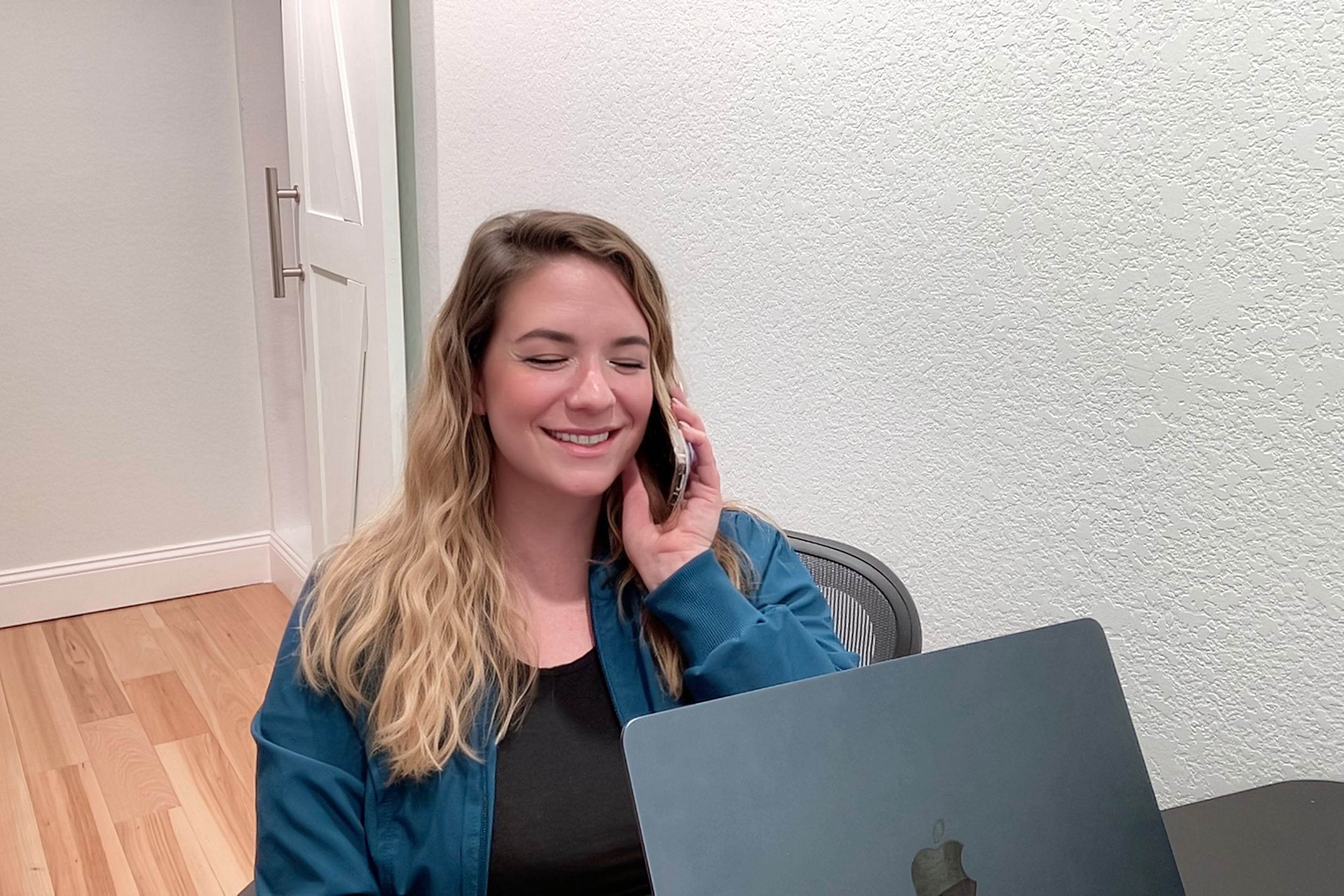 Amy Du, PMHNP, speaking with a client on a virtual appointment using her cellphone, sitting at a desk with a laptop, in a room with light-colored walls and wooden flooring.