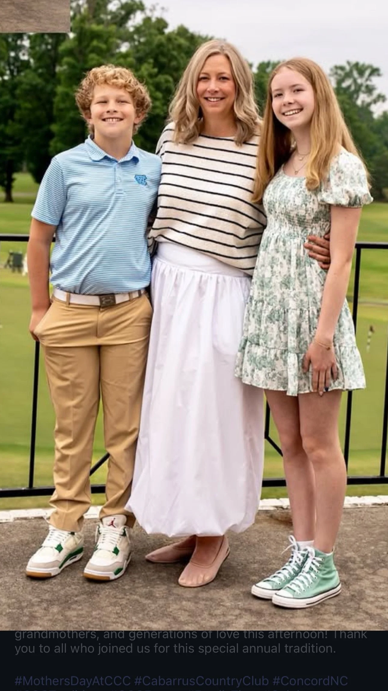 A woman with blonde wavy hair and light skin, wearing a striped shirt and white skirt, standing between a boy with curly blond hair and a girl with straight red hair, both with light skin, on a golf course with trees in the background.