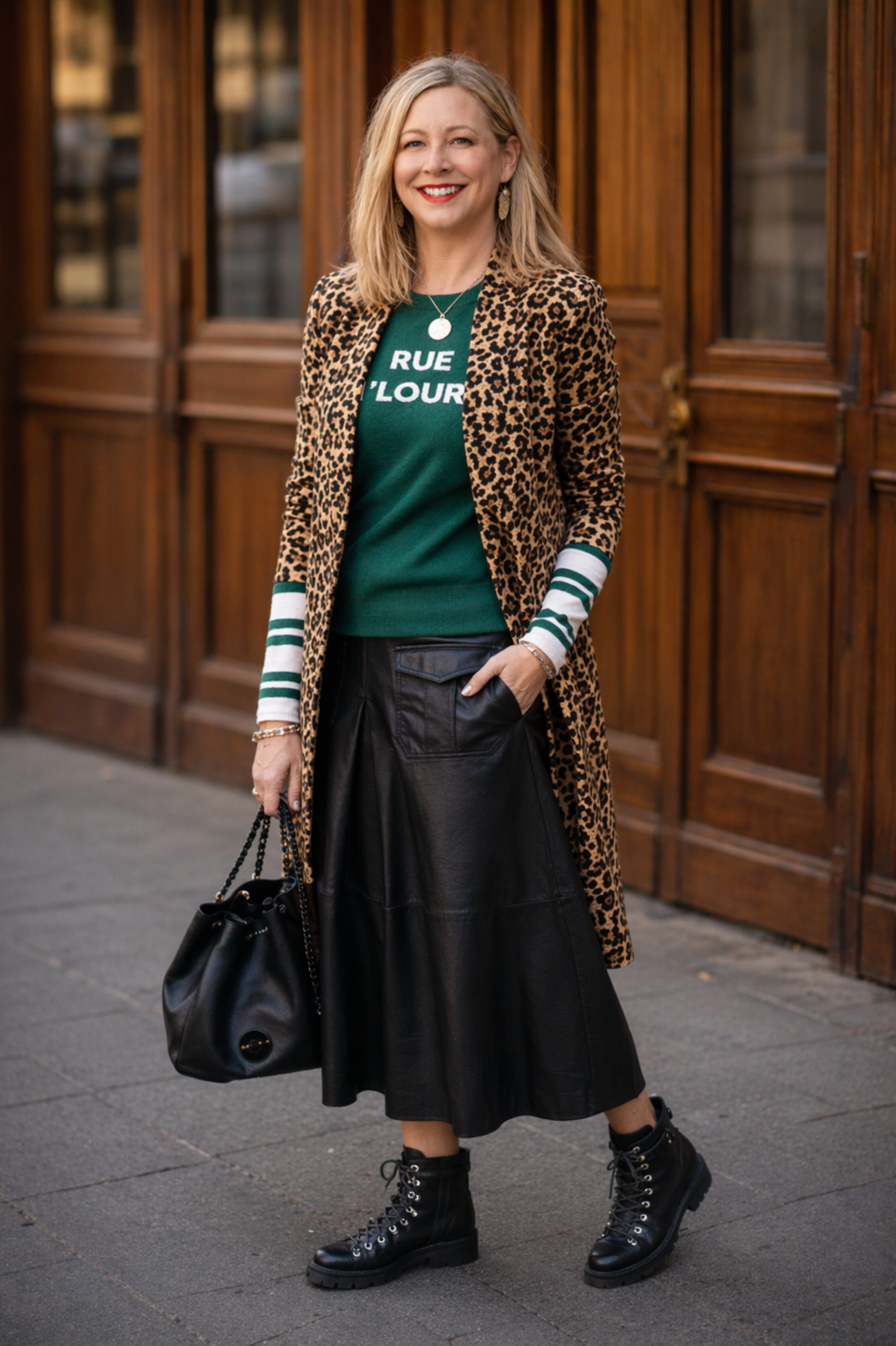 A woman standing on a city sidewalk in front of a wooden storefront, smiling and wearing a leopard print coat, green sweater with white lettering, black leather skirt, combat boots, and carrying a black purse.