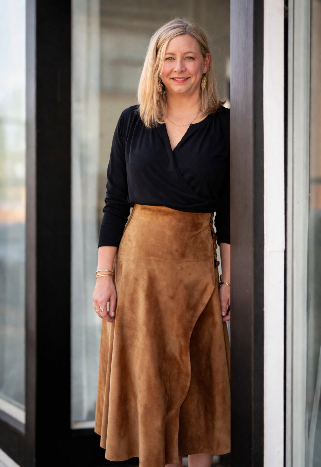 A woman with shoulder-length blonde hair, wearing a black wrap top and a brown suede skirt with side buttons, standing outside near glass and wooden panels, smiling at the camera.