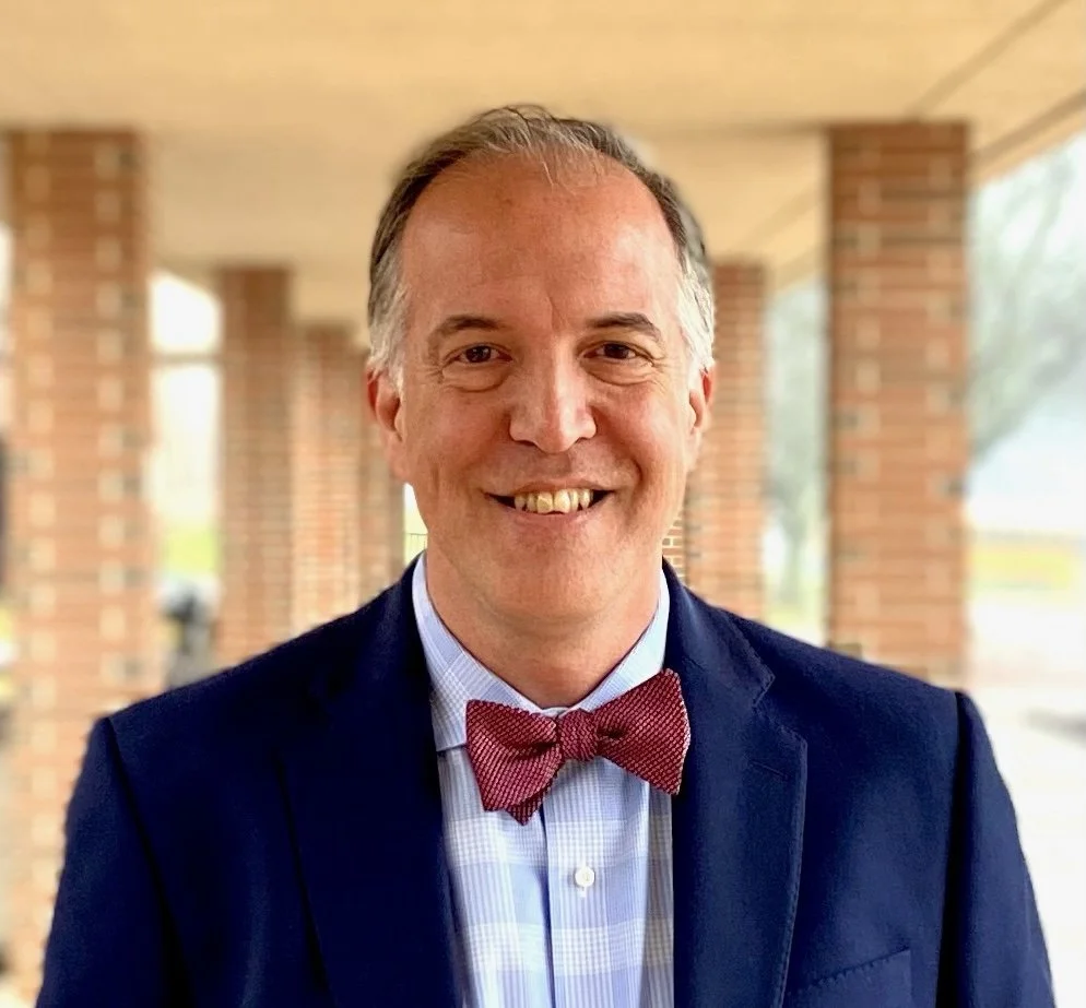 A smiling man in a navy blazer, light blue dress shirt, and red bow tie standing outside near brick columns.