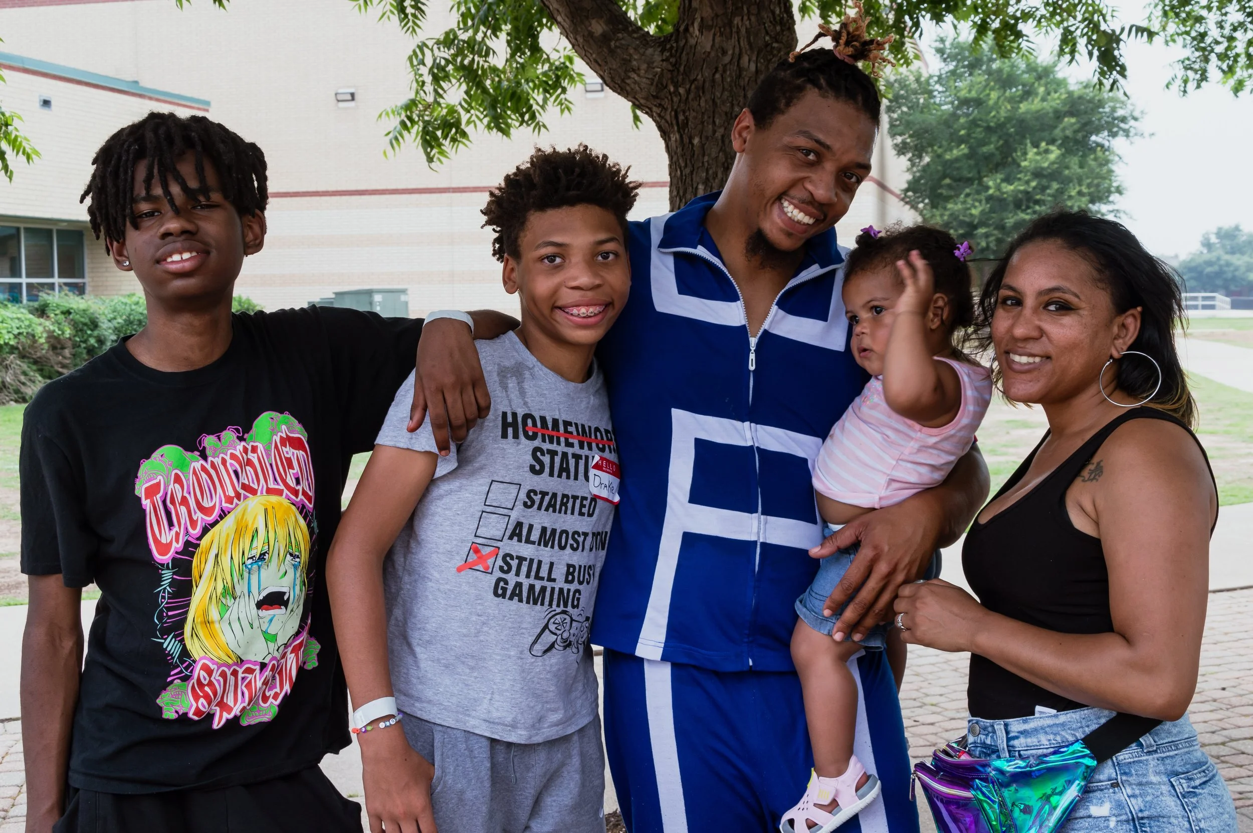 A family of five people smiling outdoors, standing under a tree, with a school building in the background. The father is holding a young girl, the mother is standing beside him, and two boys stand to the side.