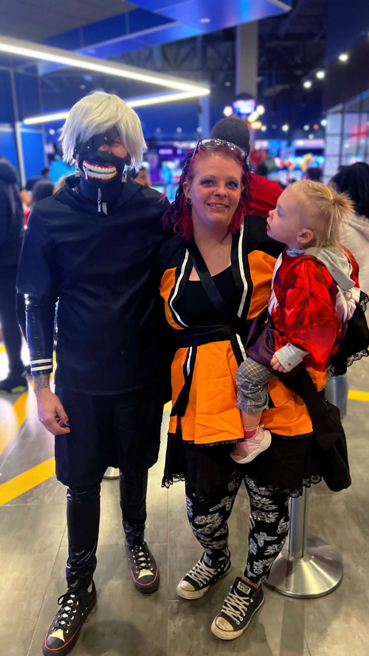 Three people standing in an indoor arcade or entertainment center, with neon lights and game machines in the background. One person is dressed as a masked character with white hair, black clothing, and a mask with a wide, painted smile. The second pe