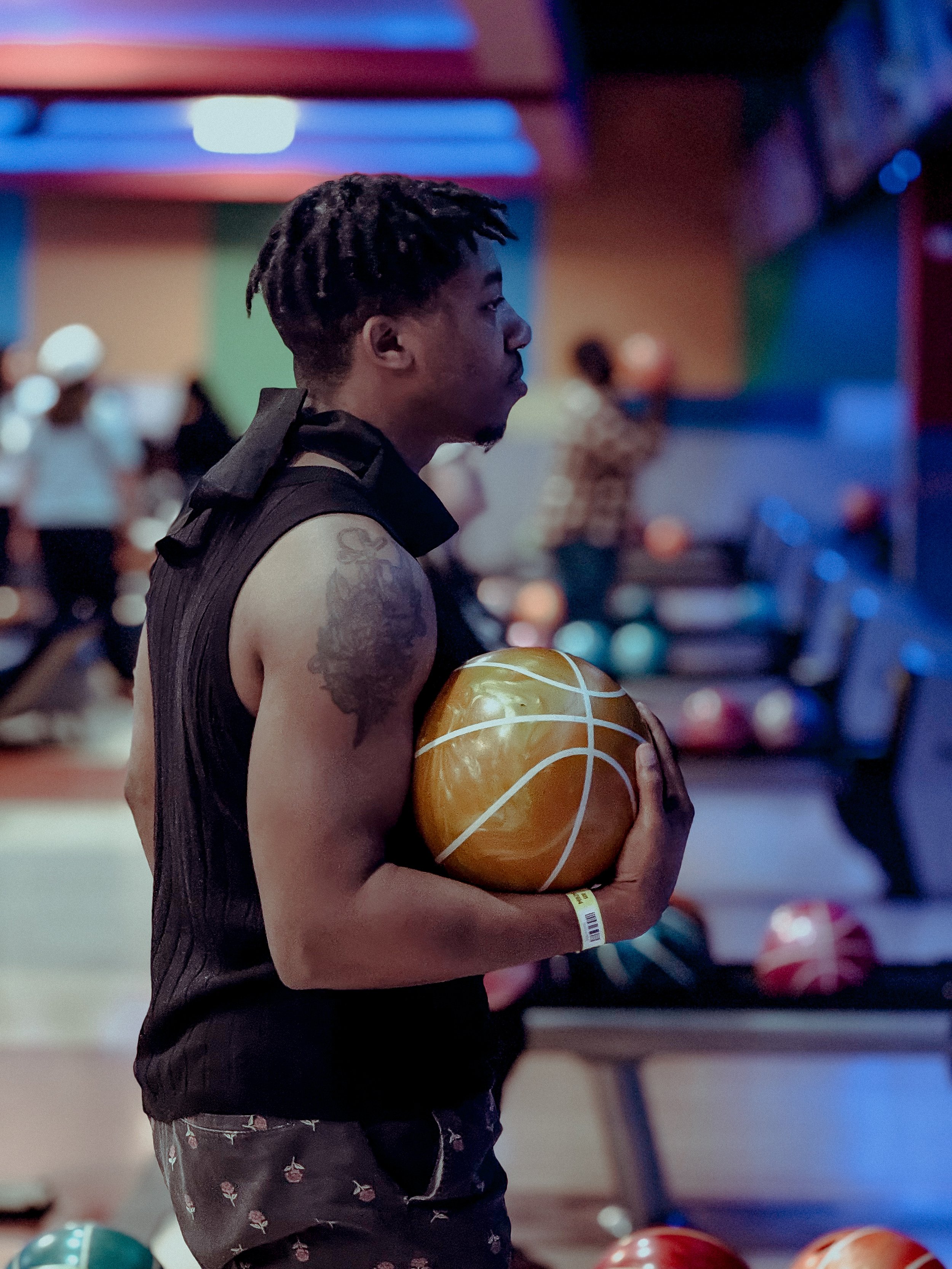 A young man holding a golden basketball at a bowling alley, surrounded by bowling balls and other people in the background.