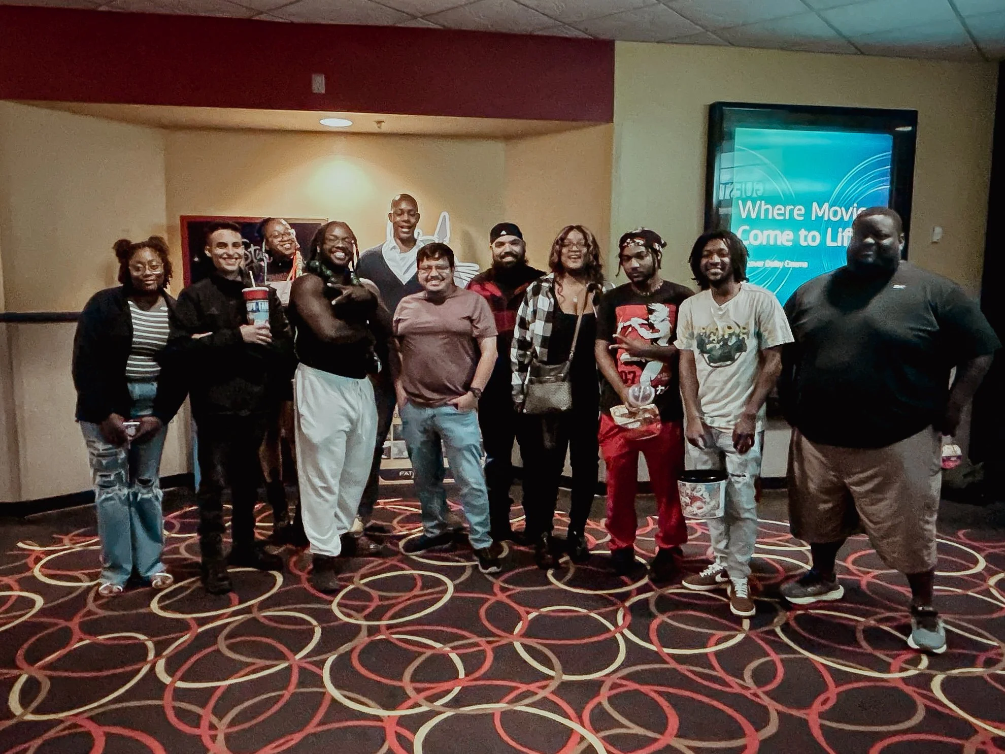 Group of eleven diverse people posing at a movie theater, standing on a patterned carpet in front of a wall with a poster. Some are smiling, holding drinks or bags, and wearing casual clothing.
