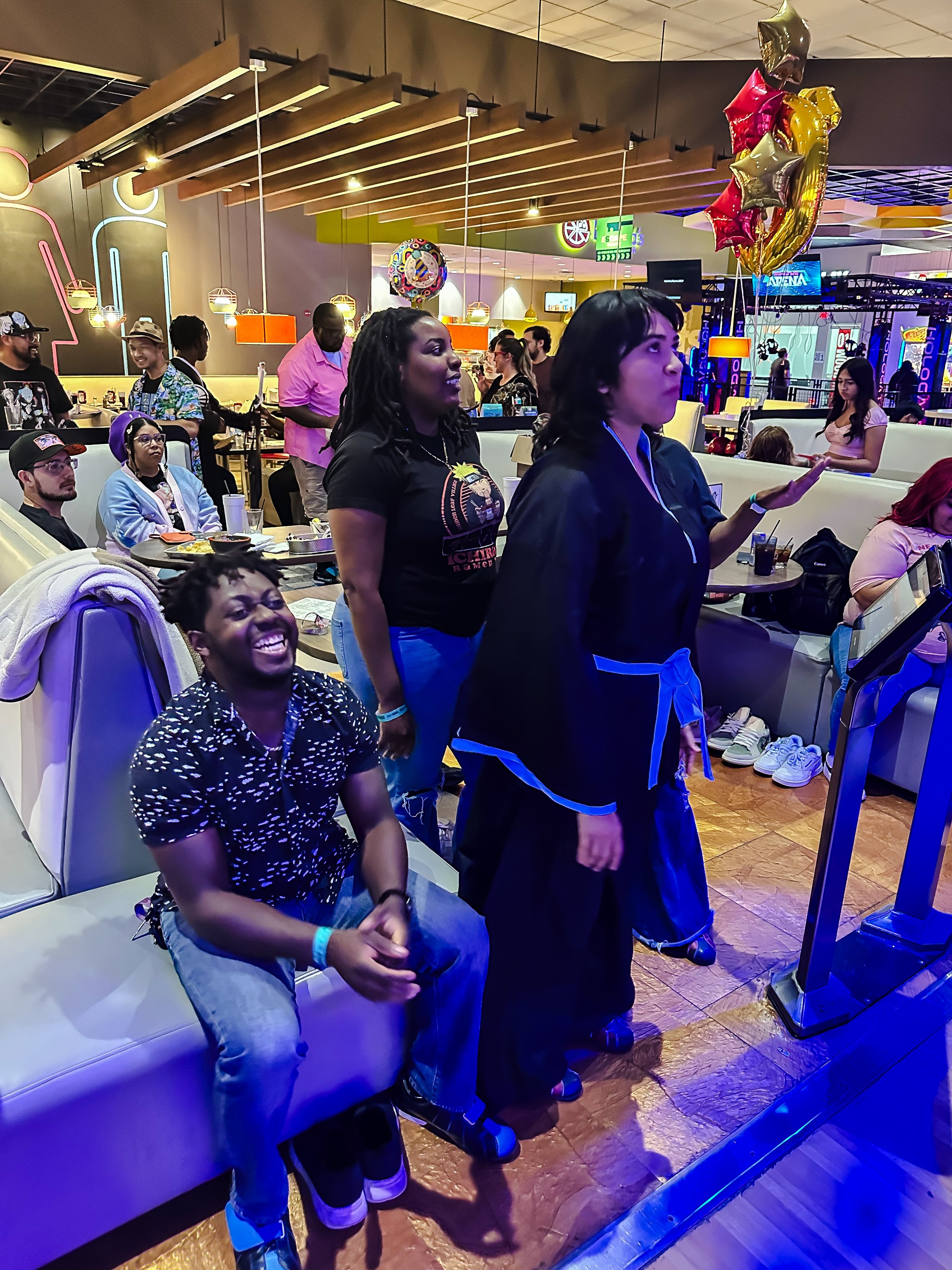 People at a bowling alley, with some seated and some standing in front of a bowling lane, surrounded by balloons and arcade games.