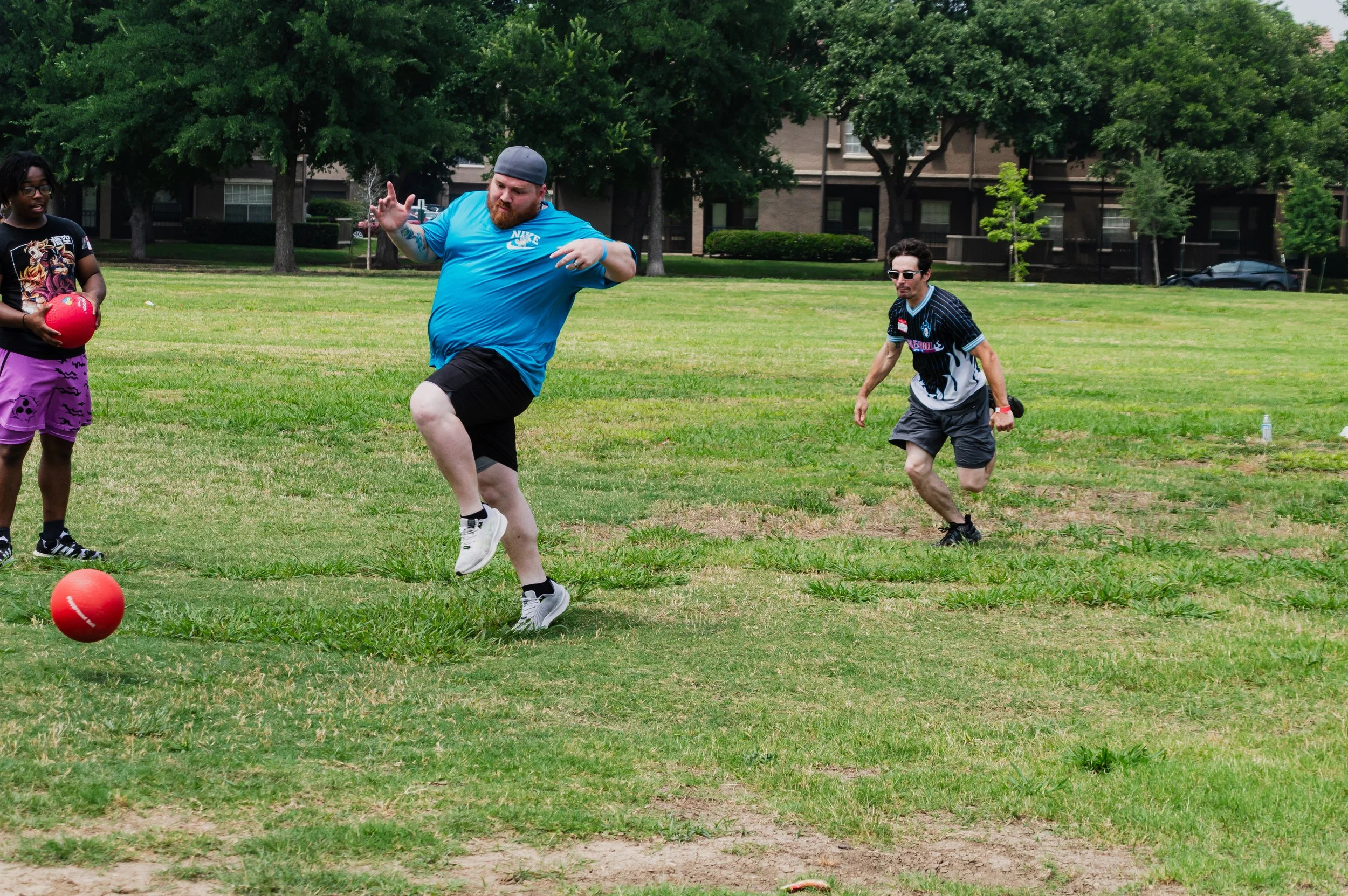 People playing dodgeball on a grassy field in a park, with trees and buildings in the background.