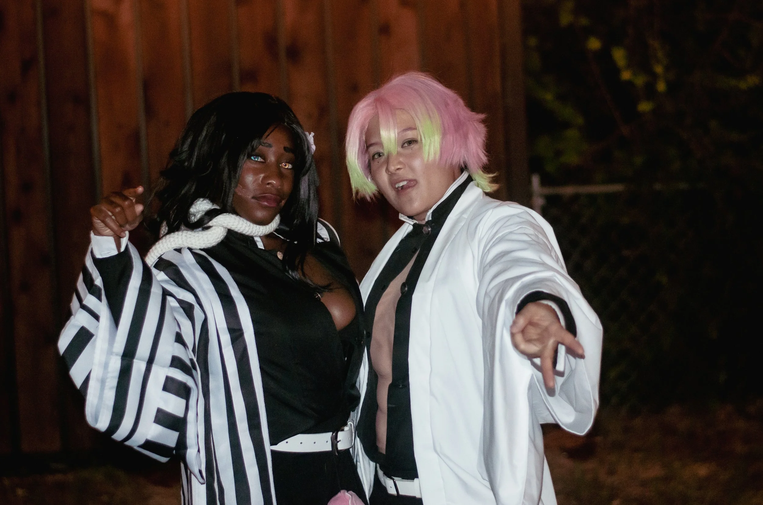 Two people dressed in cosplay costumes, one with black hair and another with pastel pink and green hair, posing outdoors at night.