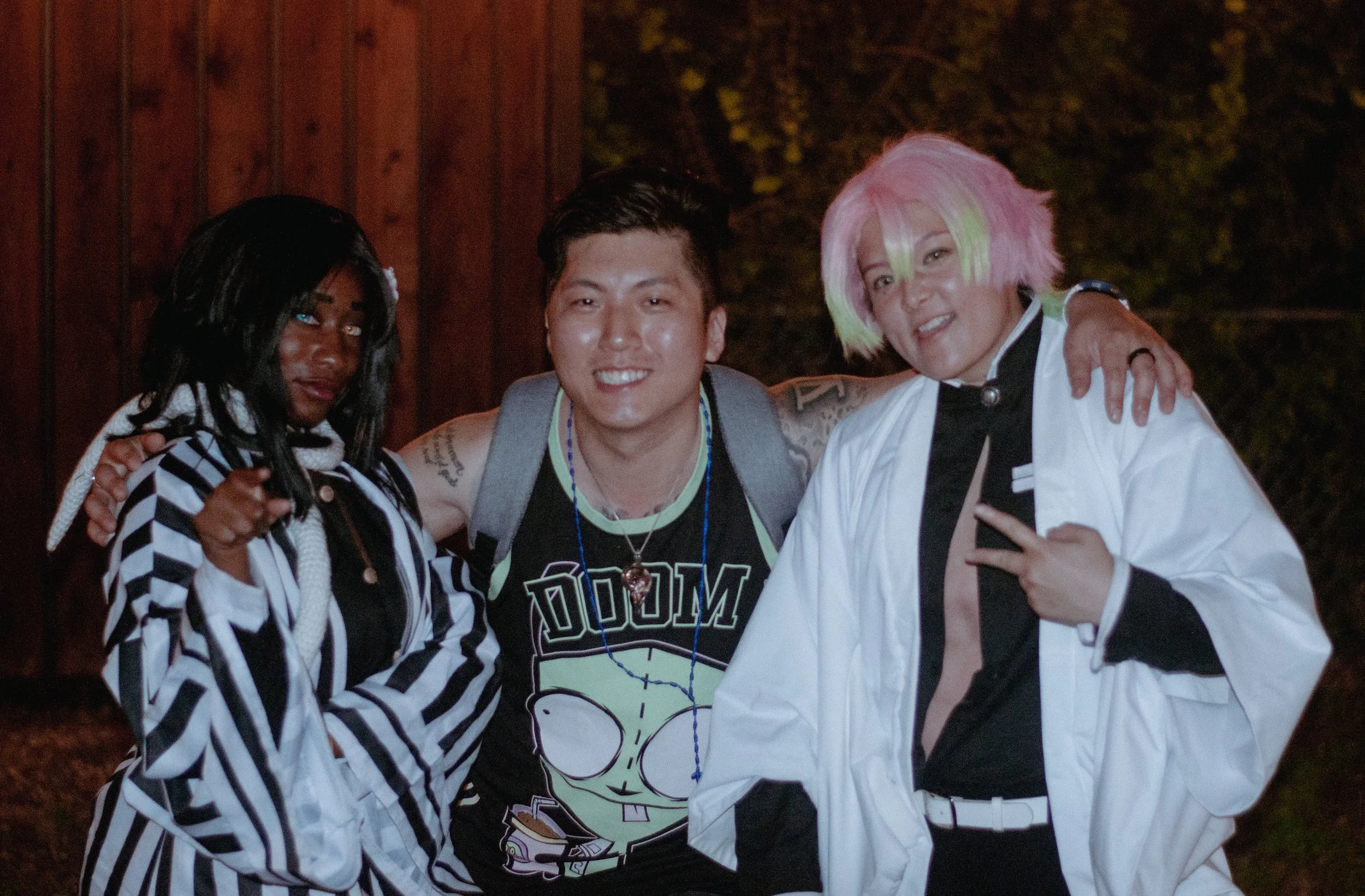 Three young adults posing together outdoors at night, with a wooden background and trees visible. The woman on the left has dark skin, black hair, and is wearing a black and white striped jacket. The man in the middle has light skin, short dark hair,