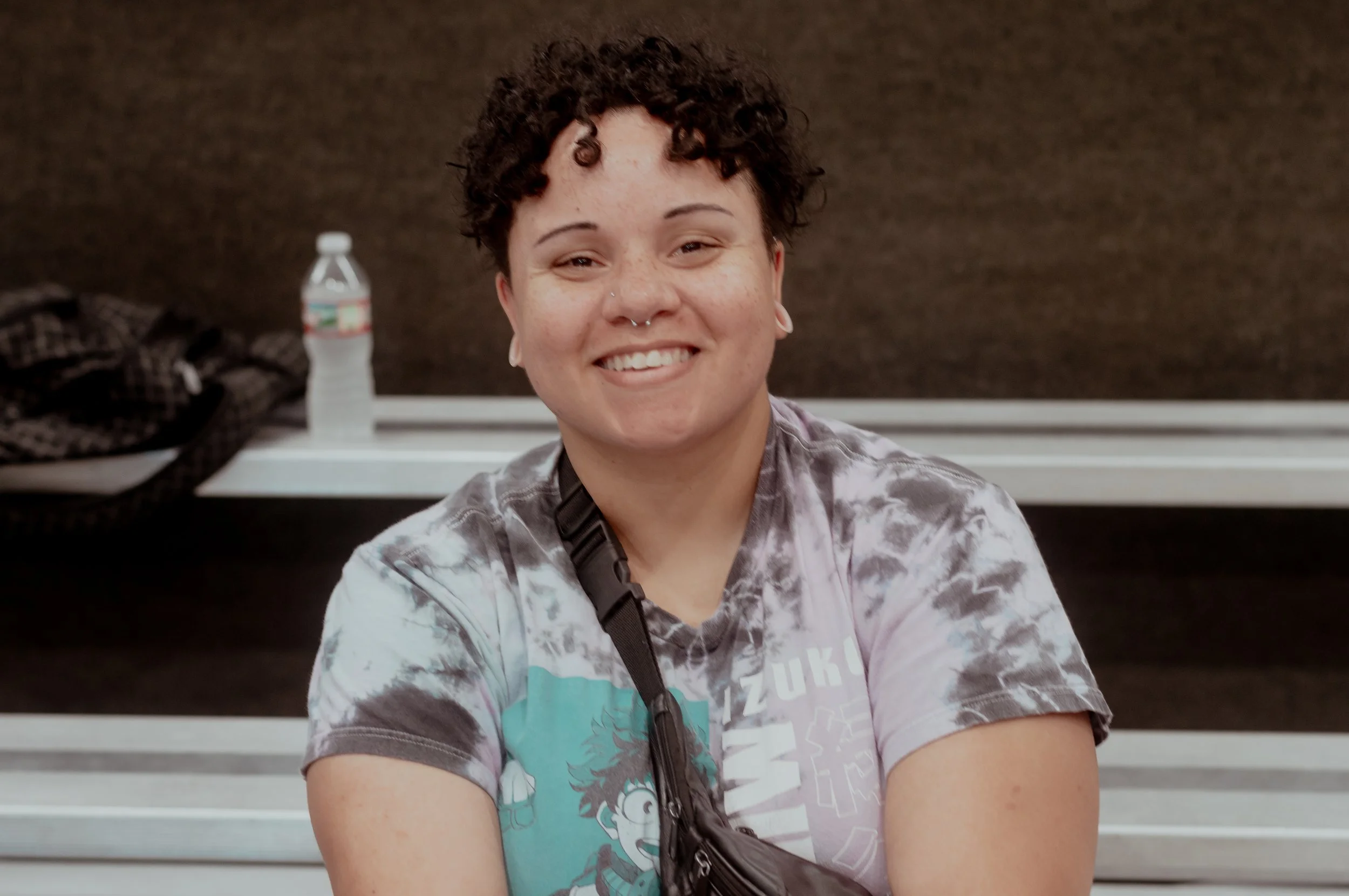 A young person with short curly hair, smiling, wearing a tie-dye shirt and a small black purse, sitting indoors against a brown background with a water bottle and some bags on a table behind them.