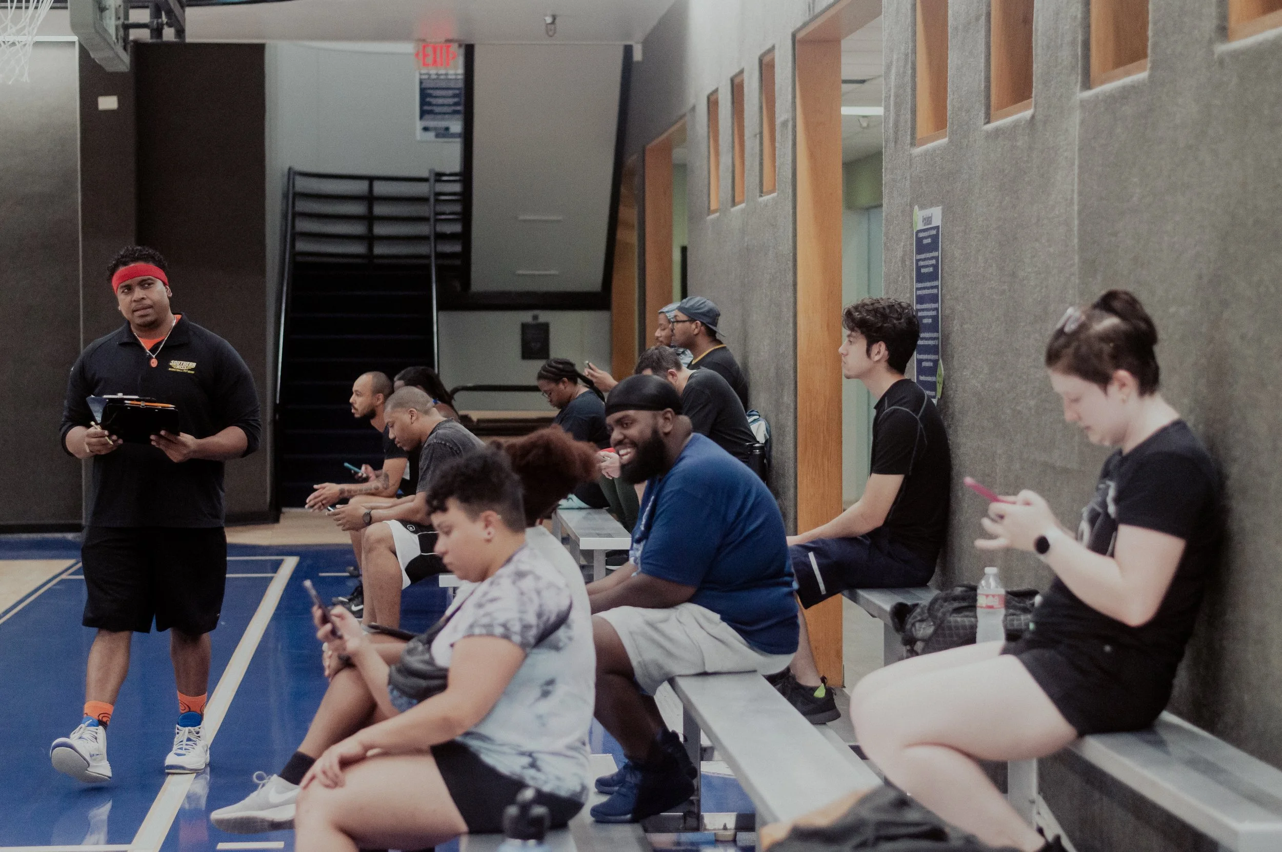 People sitting on benches in a gym or sports facility, using their phones, with a coach or instructor standing nearby holding a clipboard.