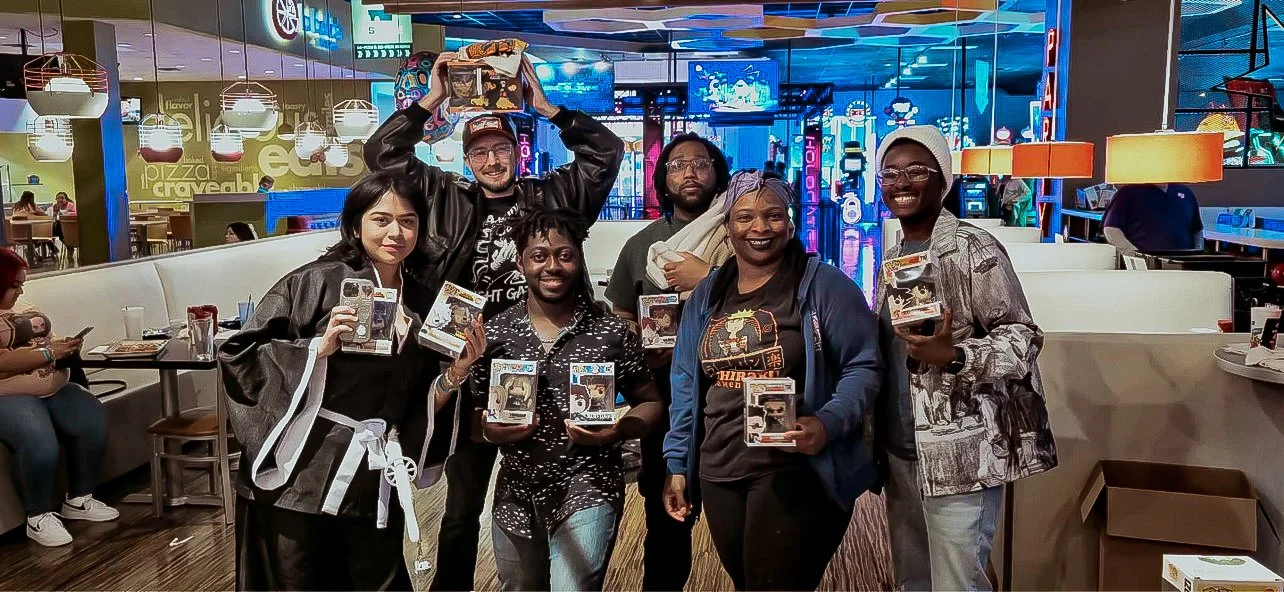 Group of seven people smiling and holding box sets of Funko Pop figurines in a lively arcade or entertainment center.