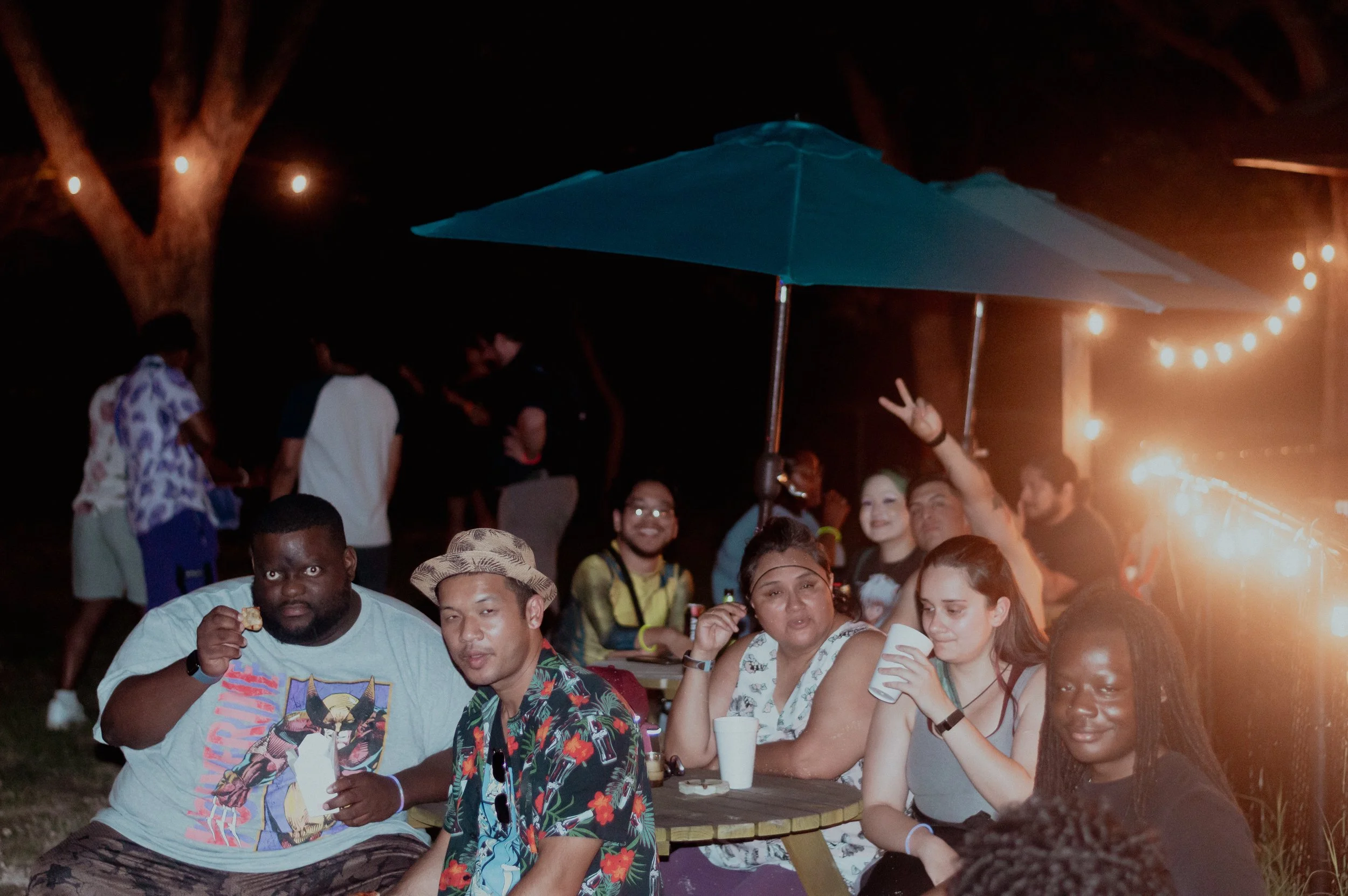 Group of people sitting around a table at night, some drinking and chatting, with string lights and a large umbrella in the background.