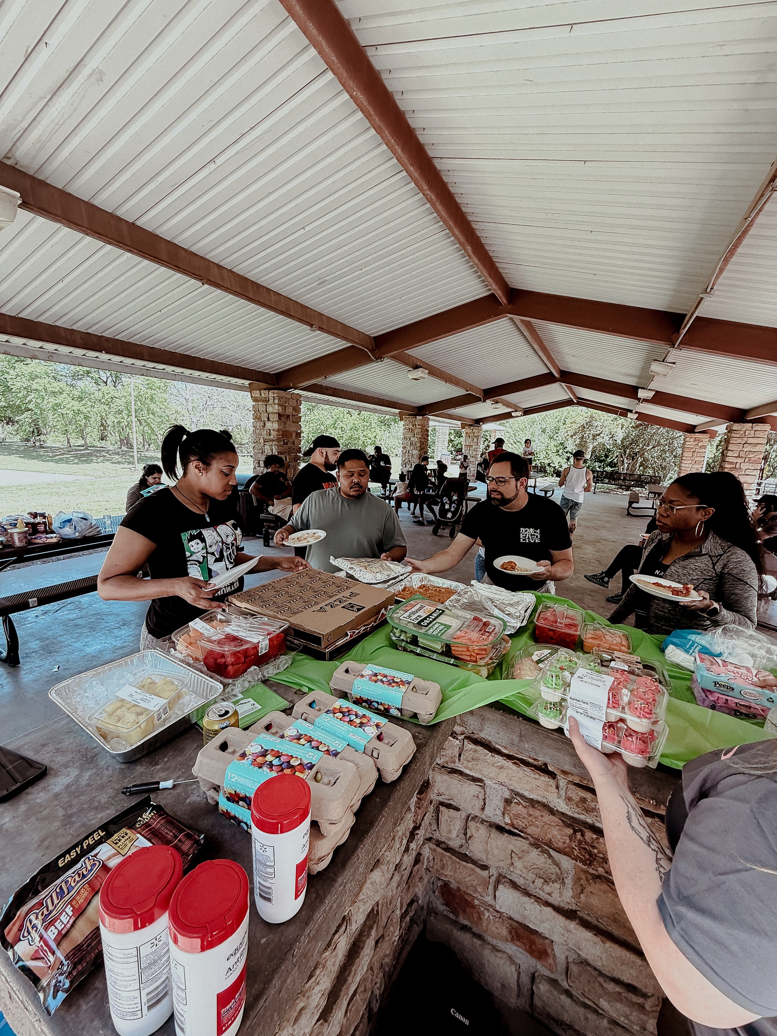A group of people at a potluck-style outdoor gathering under a pavilion with a brick counter filled with food and drinks.