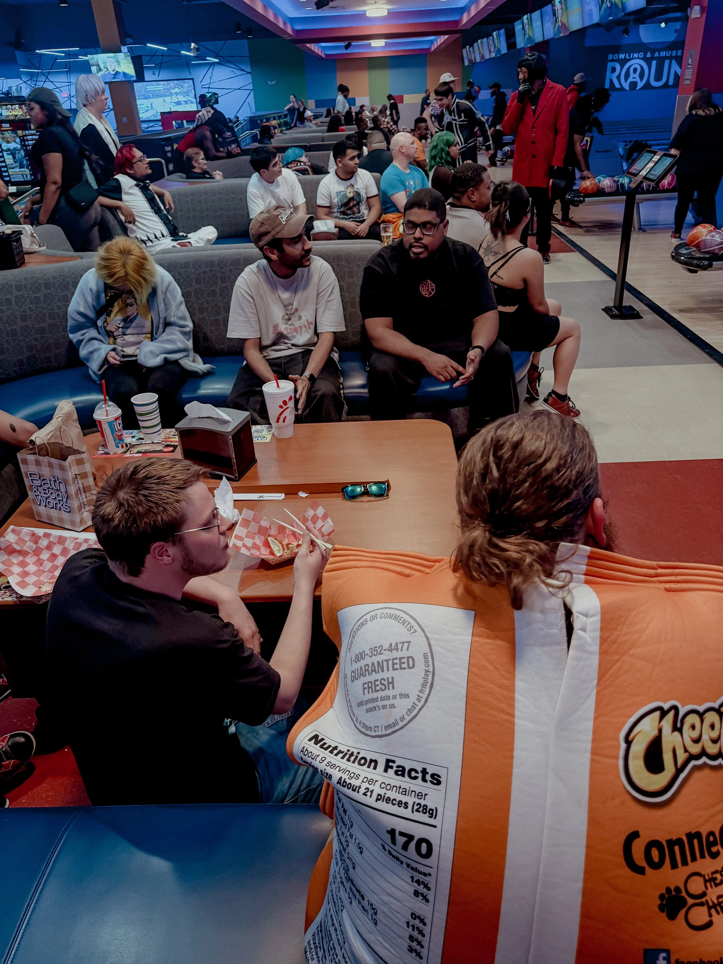People sitting and waiting at a bowling alley, with some watching a game, others eating snacks, and a man walking with a red coat in the background.