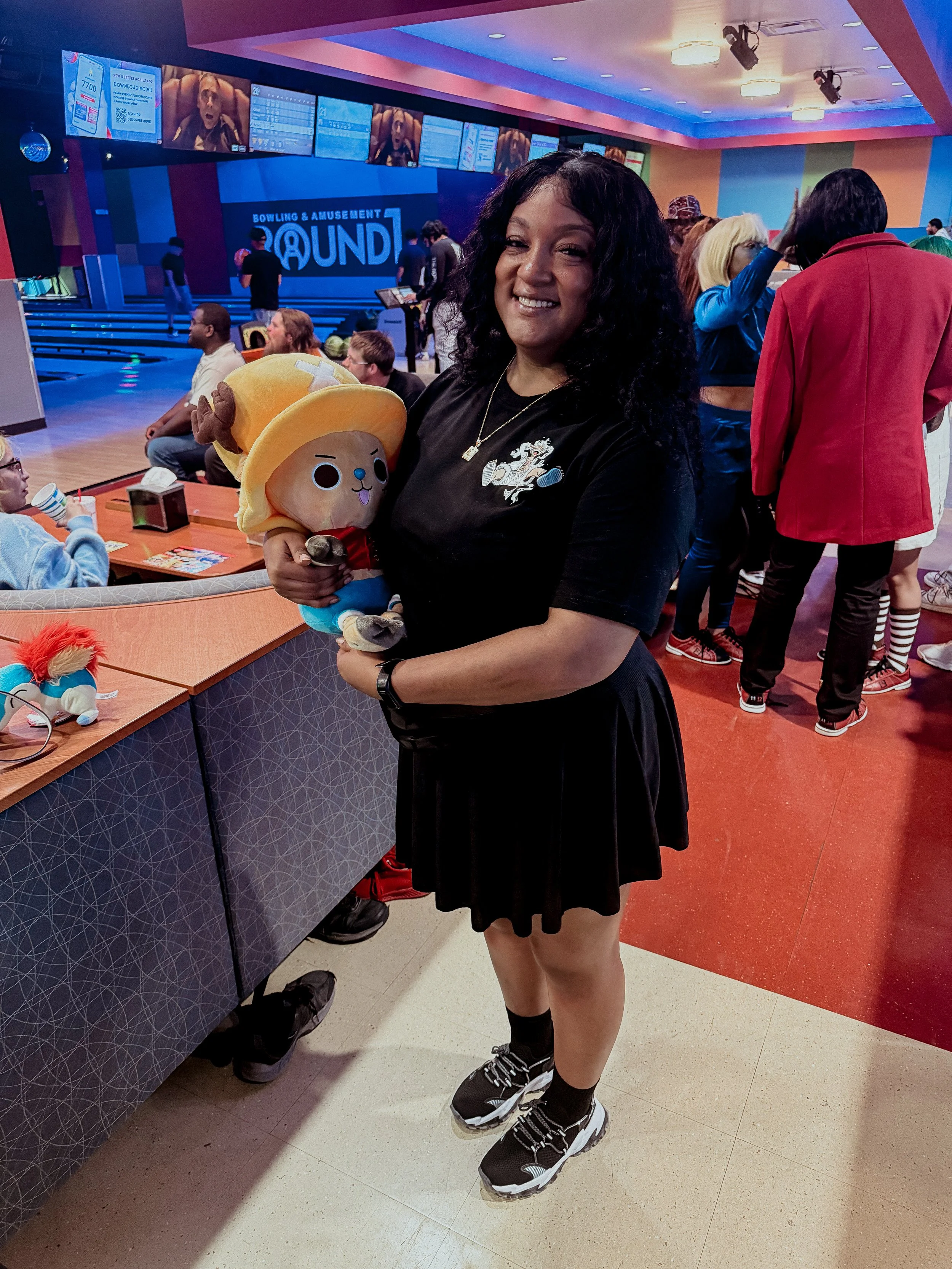 A woman with curly black hair smiling and standing in a bowling alley, holding a plush toy with a large hat. The background shows other people, some sitting and some standing, with bowling lanes and screens above showing scores.
