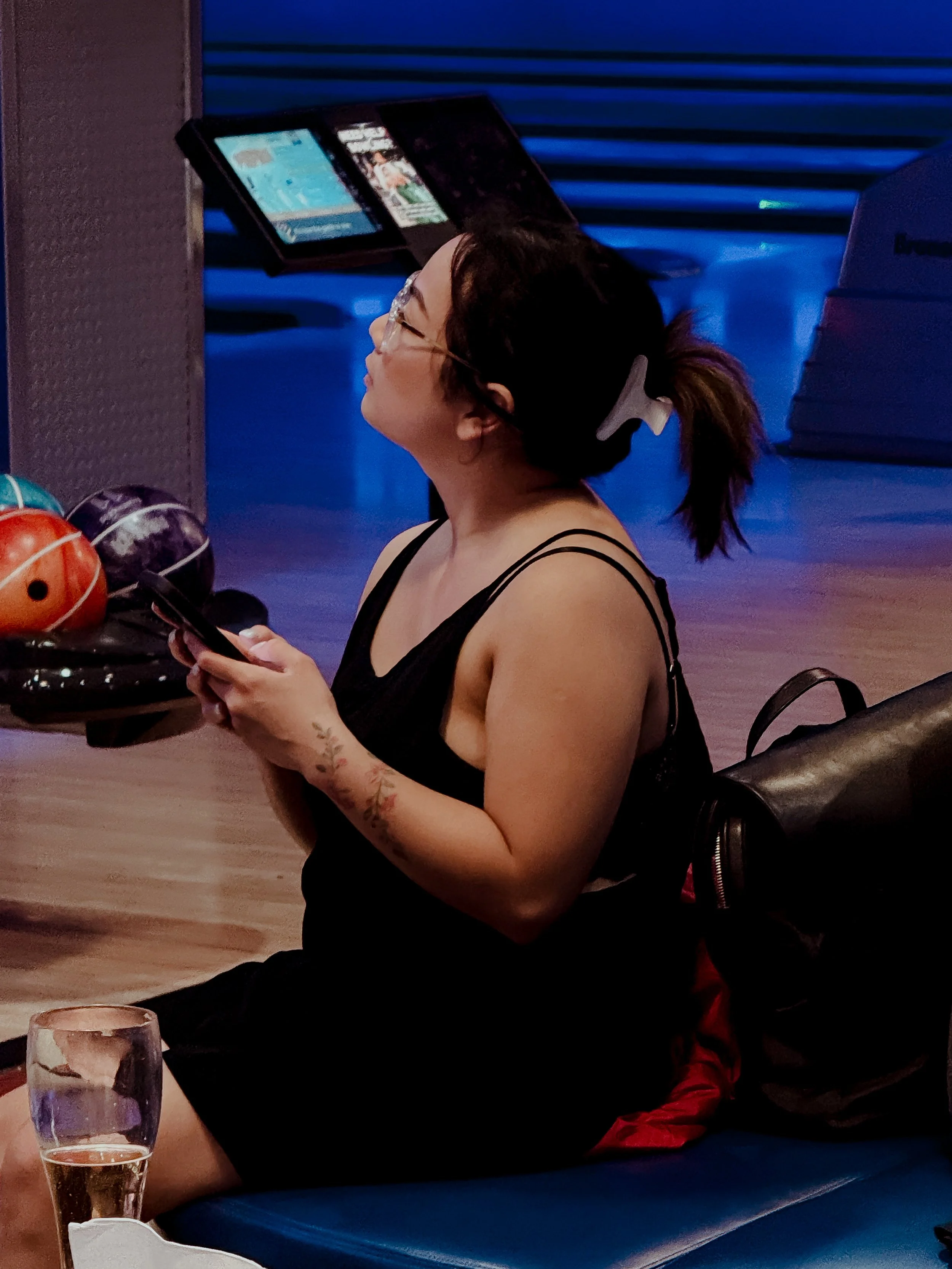 A woman with glasses and a ponytail holding a smartphone, sitting at a bowling alley with bowling balls nearby, a water glass in front, and illuminated screens in the background.
